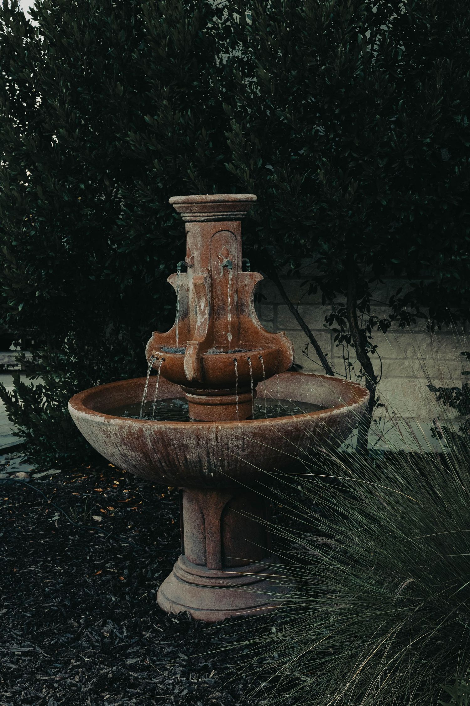 Rustic terracotta two-tier garden fountain with flowing water surrounded by lush green shrubs.