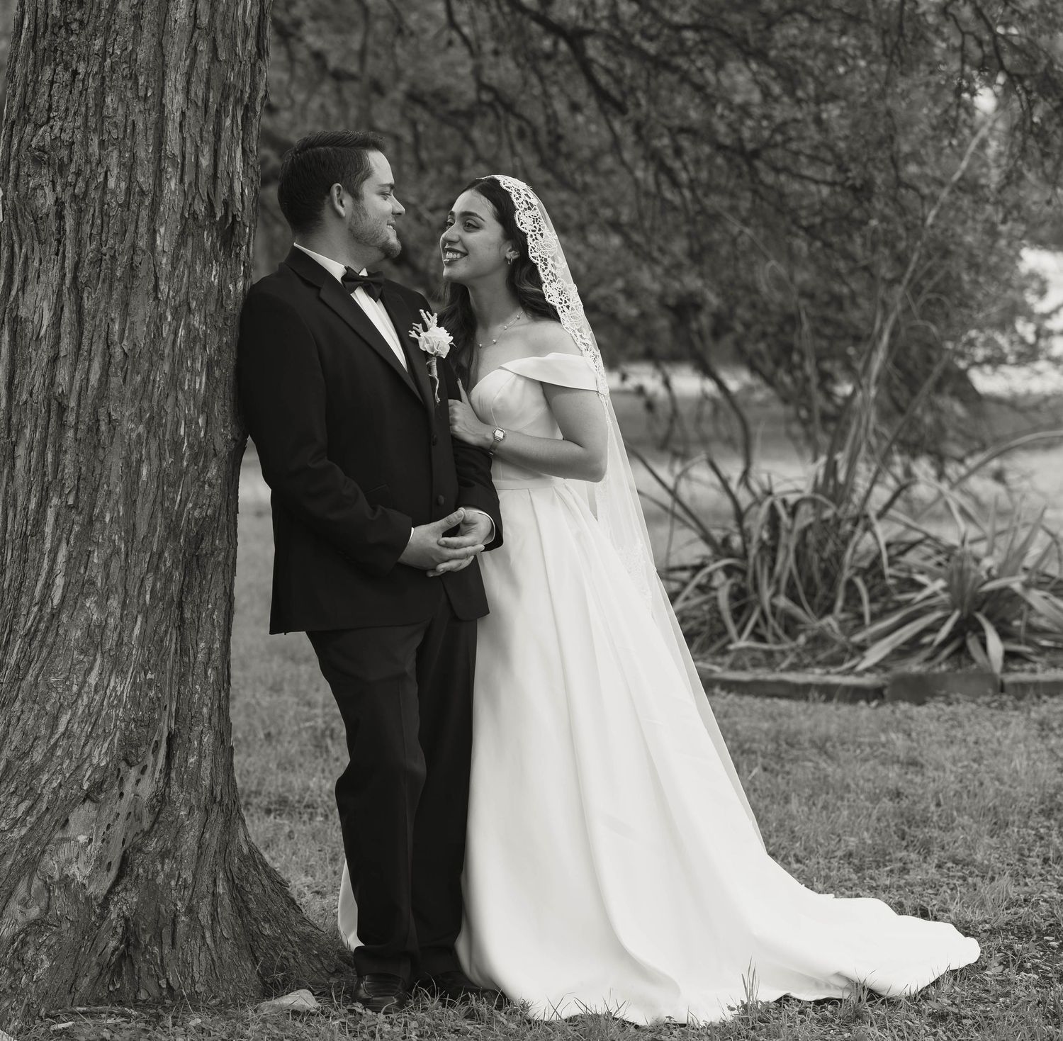 Bride and groom share a tender moment leaning against a tree in a black and white wedding portrait.