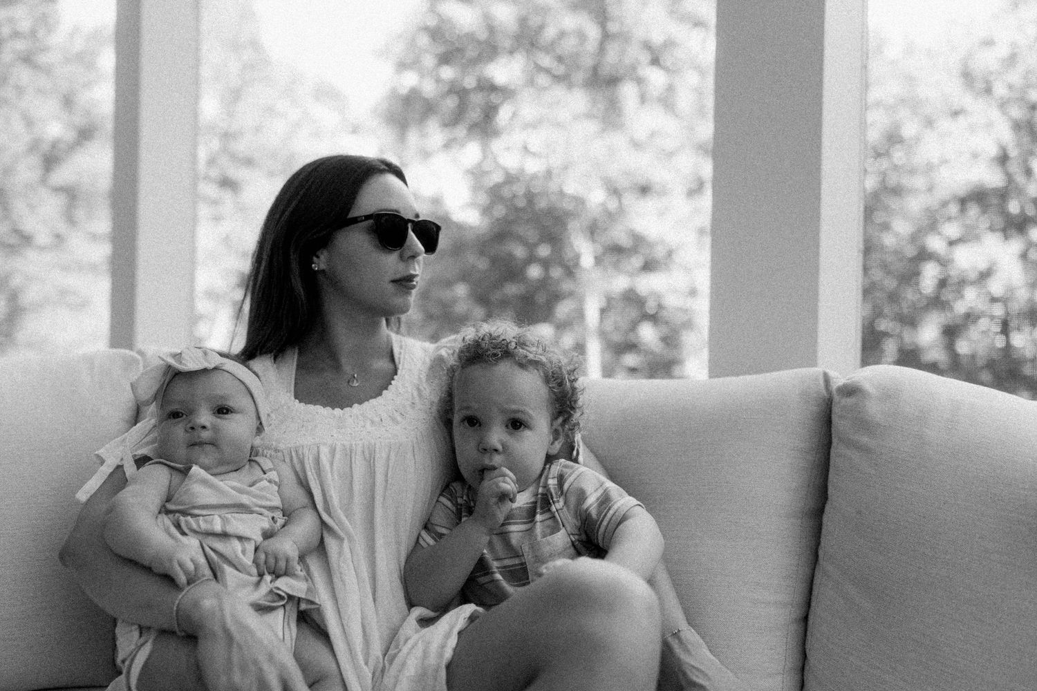 Black and white photo of a mother in sunglasses sitting on a couch with her baby and toddler by large windows.