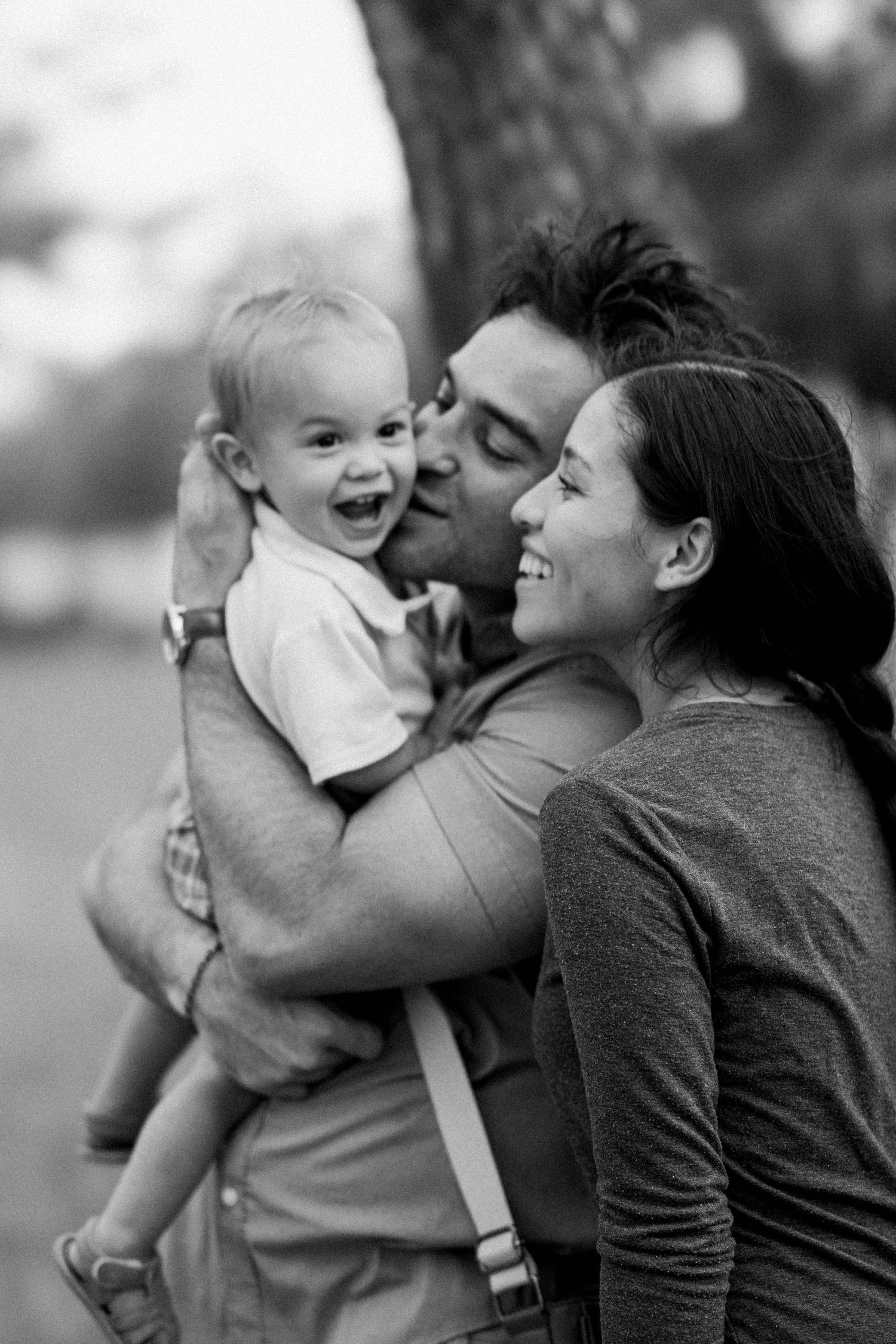 Black and white photo of happy parents kissing their laughing baby outdoors.