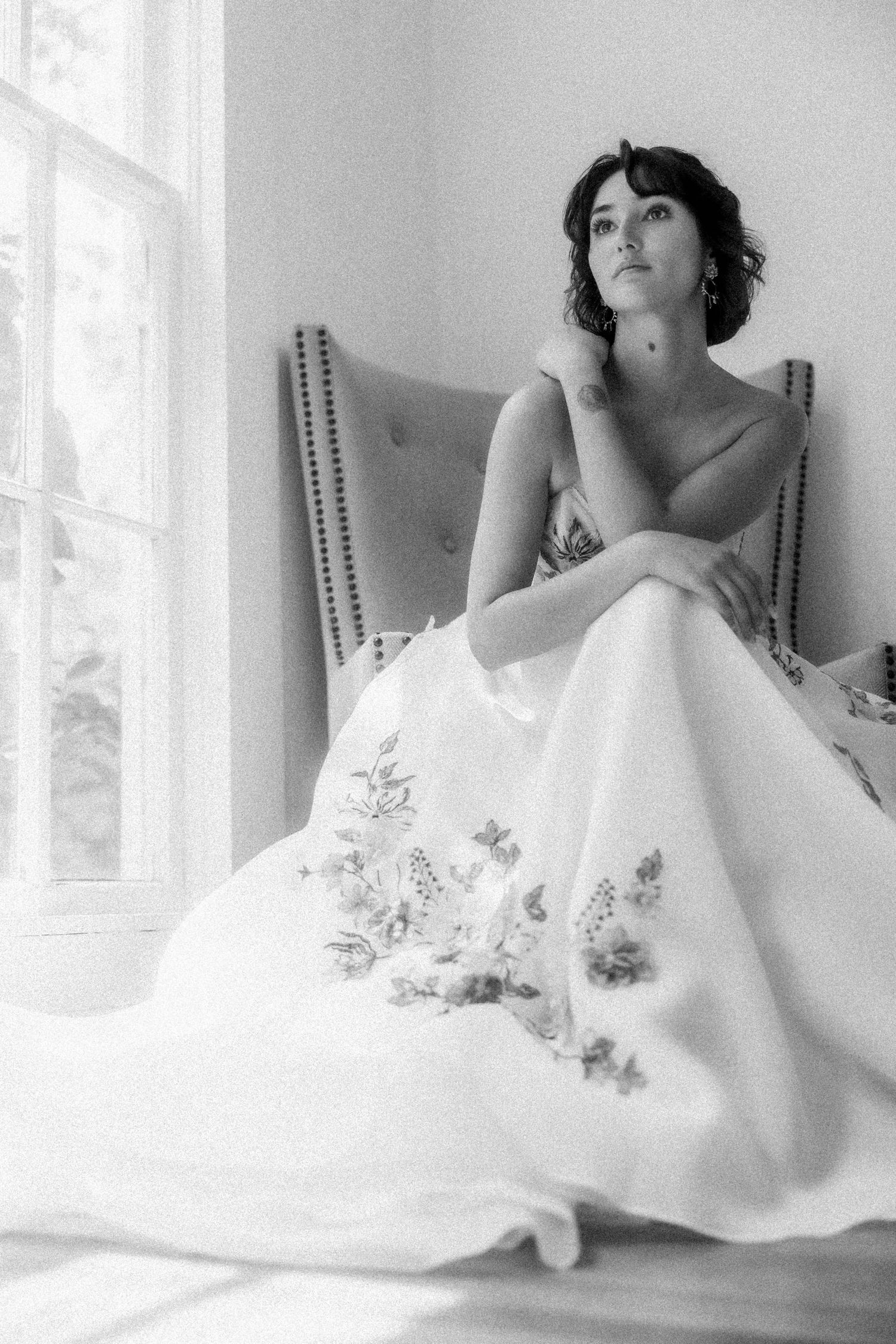 Black and white photo of a bride in a floral embroidered gown seated by a window.