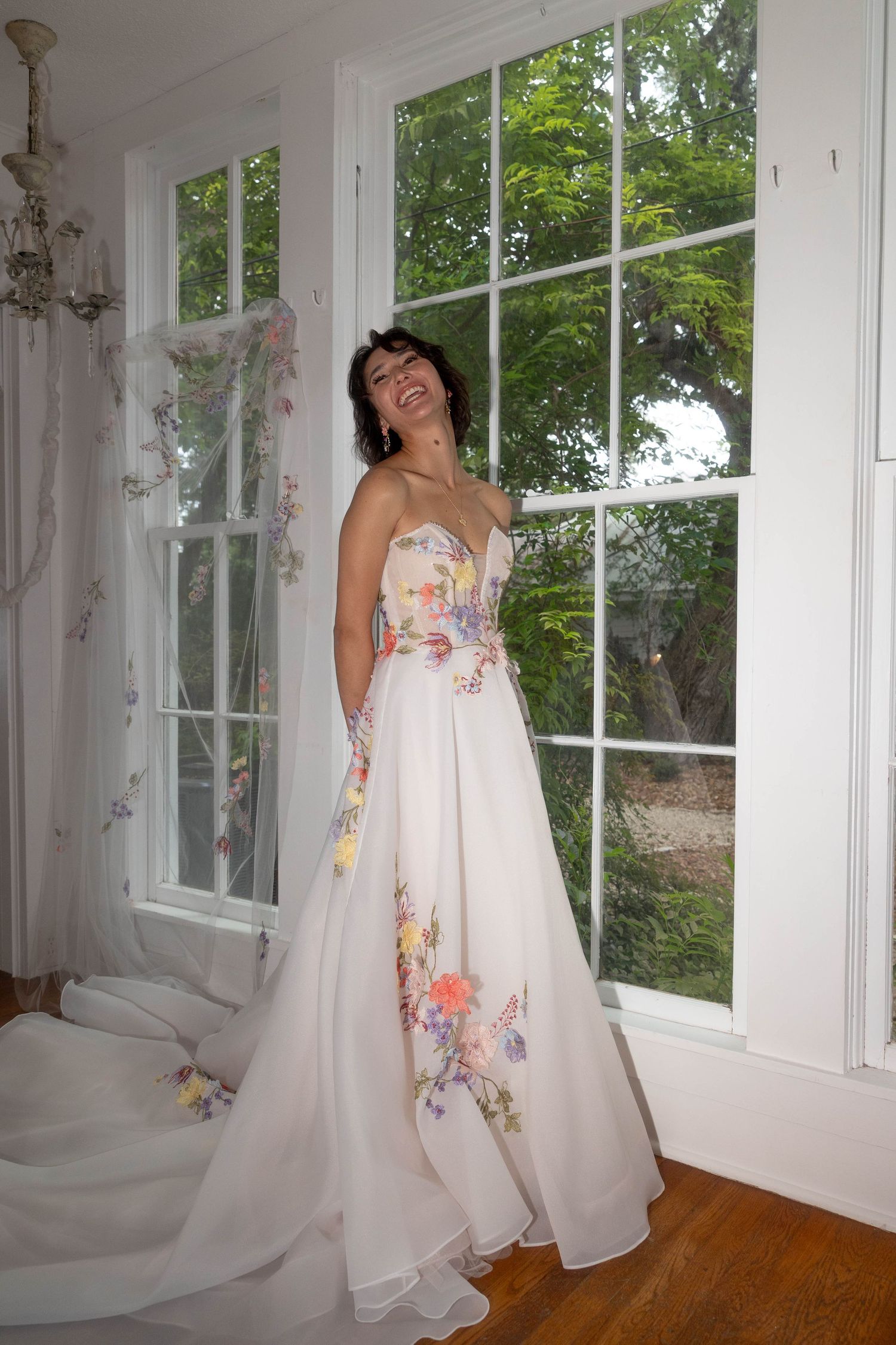 Bride laughing joyfully in strapless floral wedding dress near bright windows.
