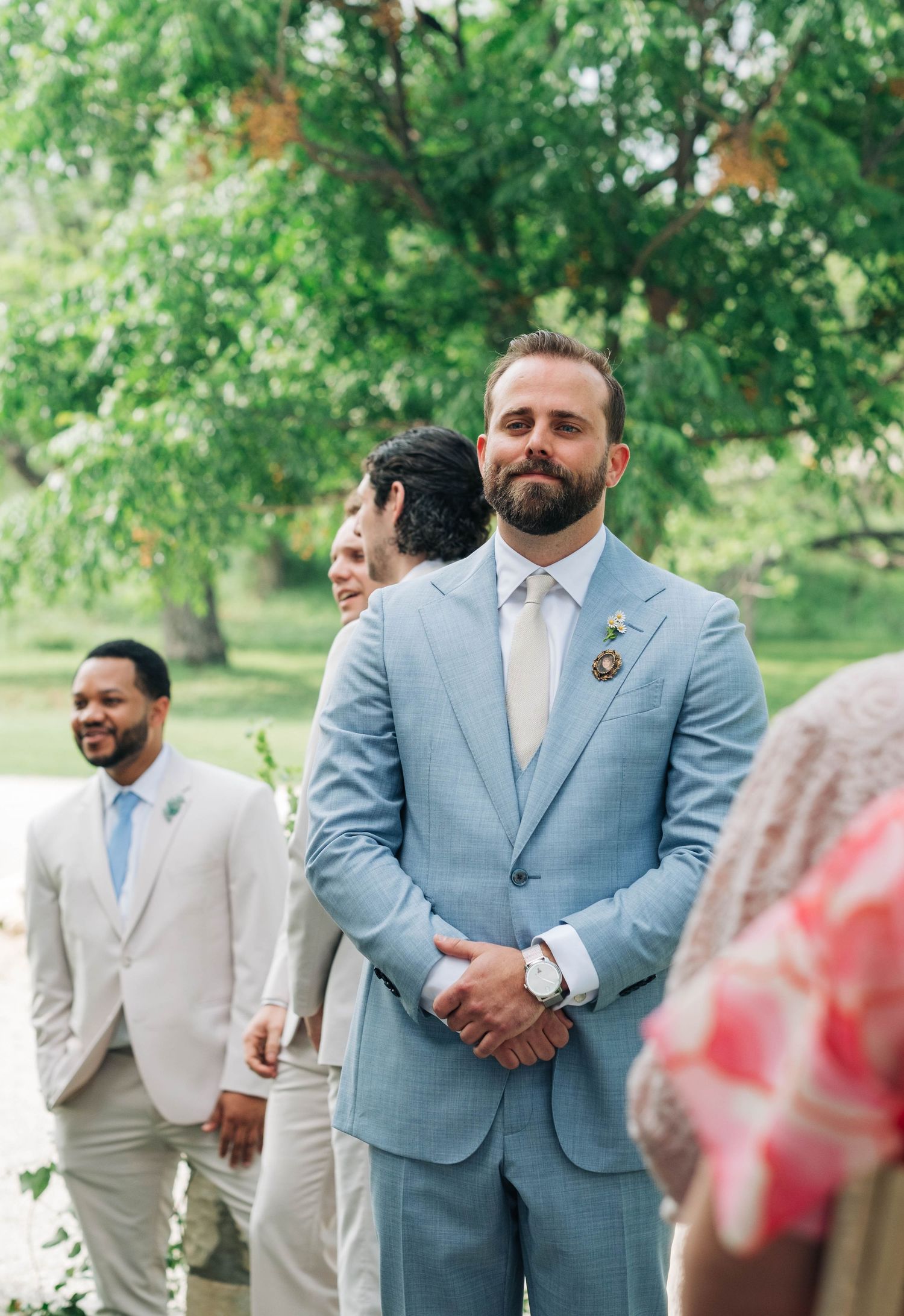 Groom in a light blue suit with a floral boutonniere waiting at the altar during an outdoor ceremony.
