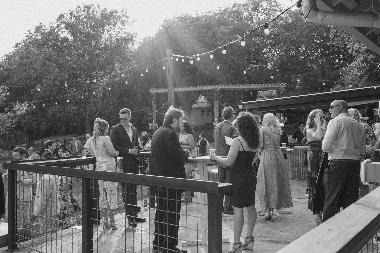 Black and white photo of elegantly dressed wedding guests mingling on an outdoor patio with string lights.