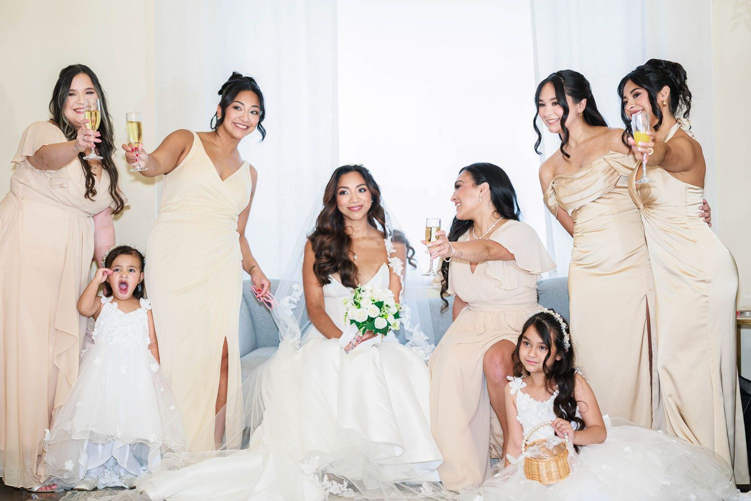 Bride and bridesmaids in champagne gowns celebrate with flower girls and sparkling wine.