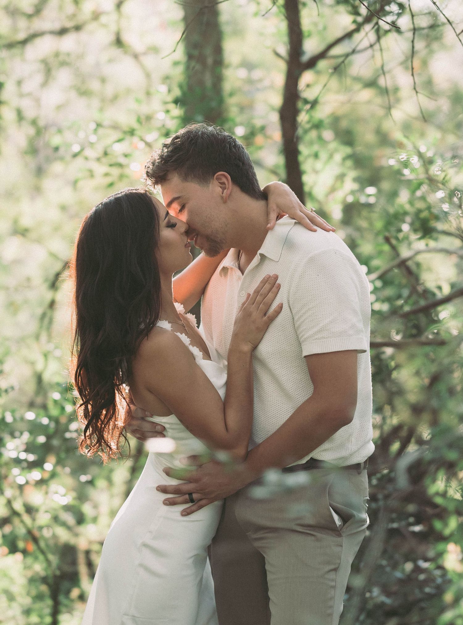 Couple sharing a romantic kiss while embracing in a sunlit forest setting.