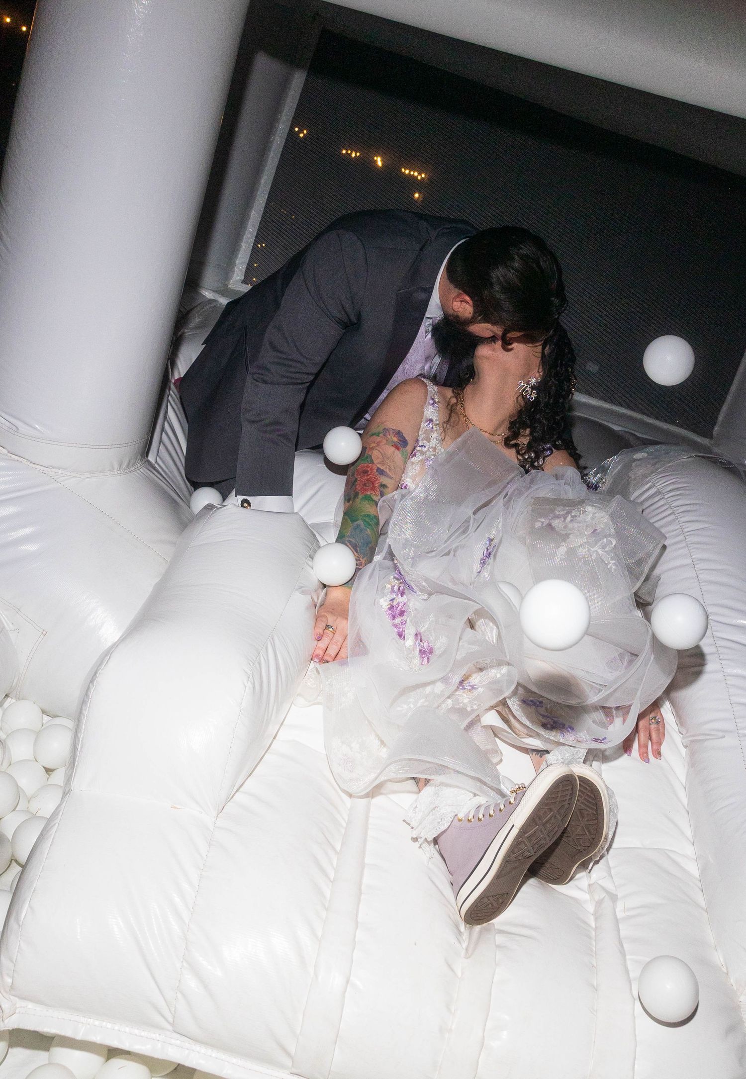 Bearded groom leans down to kiss tattooed bride lying in white inflatable bounce house at wedding reception.
