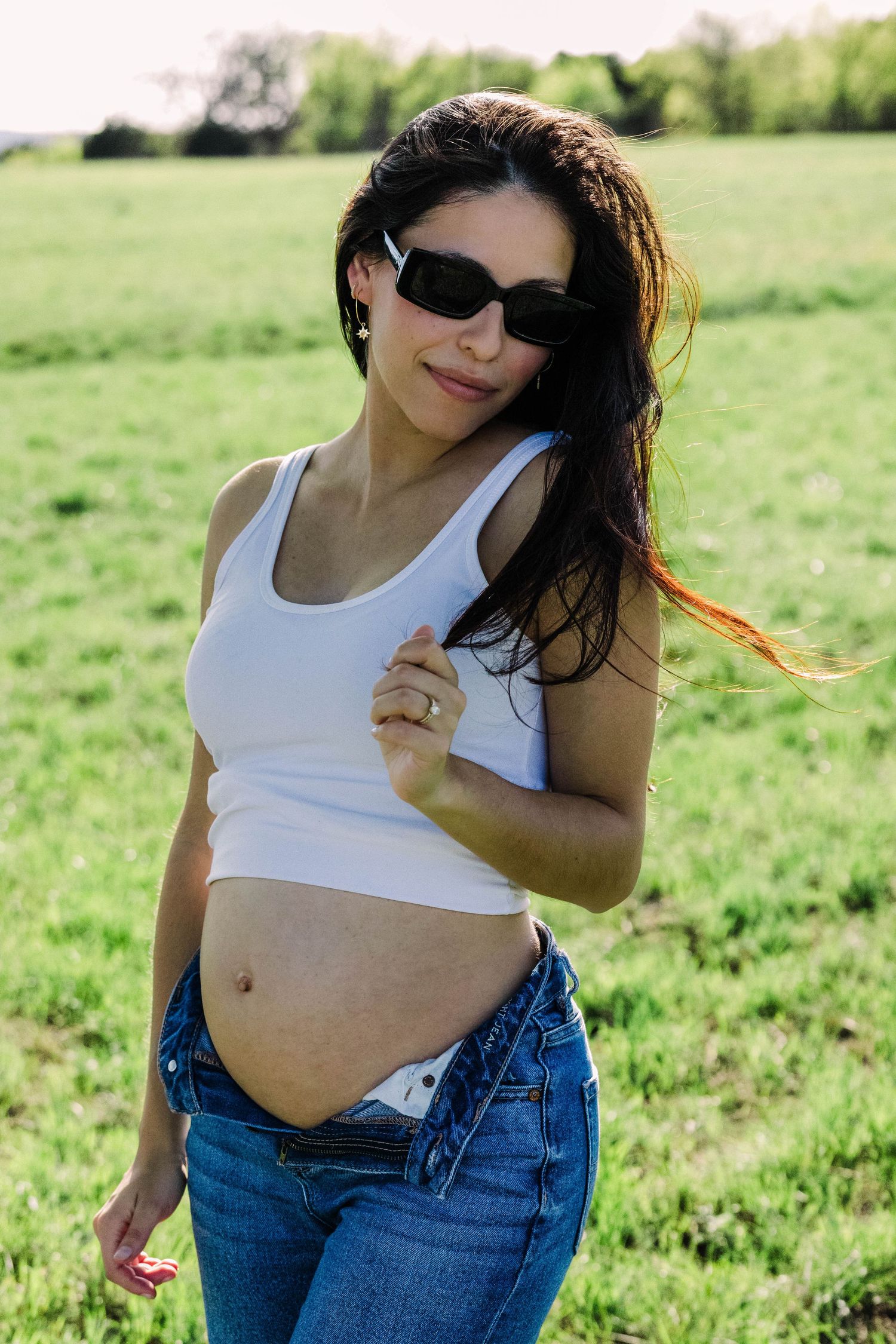 Pregnant woman in white tank top and unbuttoned jeans posing in sunny green field outdoors.
