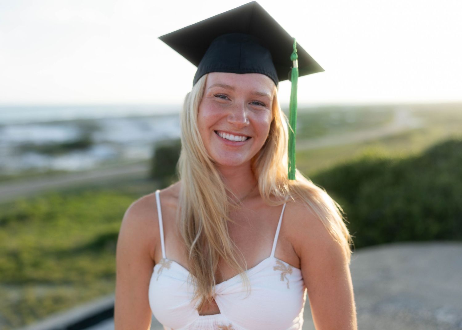 Smiling blonde woman wearing black graduation cap near coastal outdoor setting.