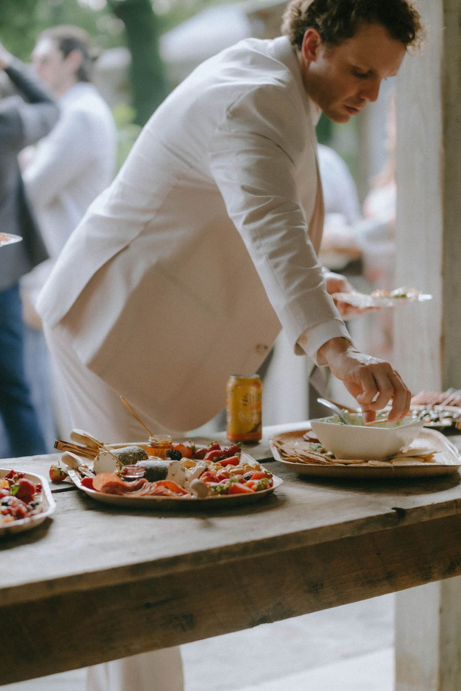 Elegantly dressed man in white suit serves himself from a wedding cocktail hour charcuterie spread.