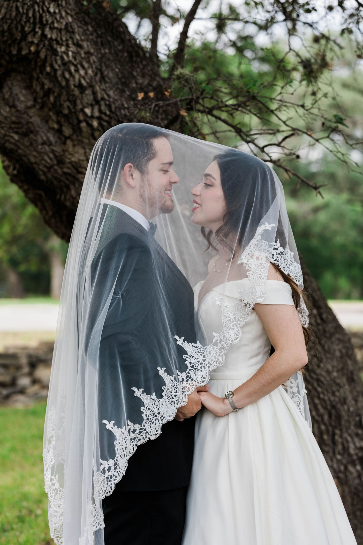 Bride and groom smile at each other under a lace-trimmed wedding veil beside a large tree outdoors.