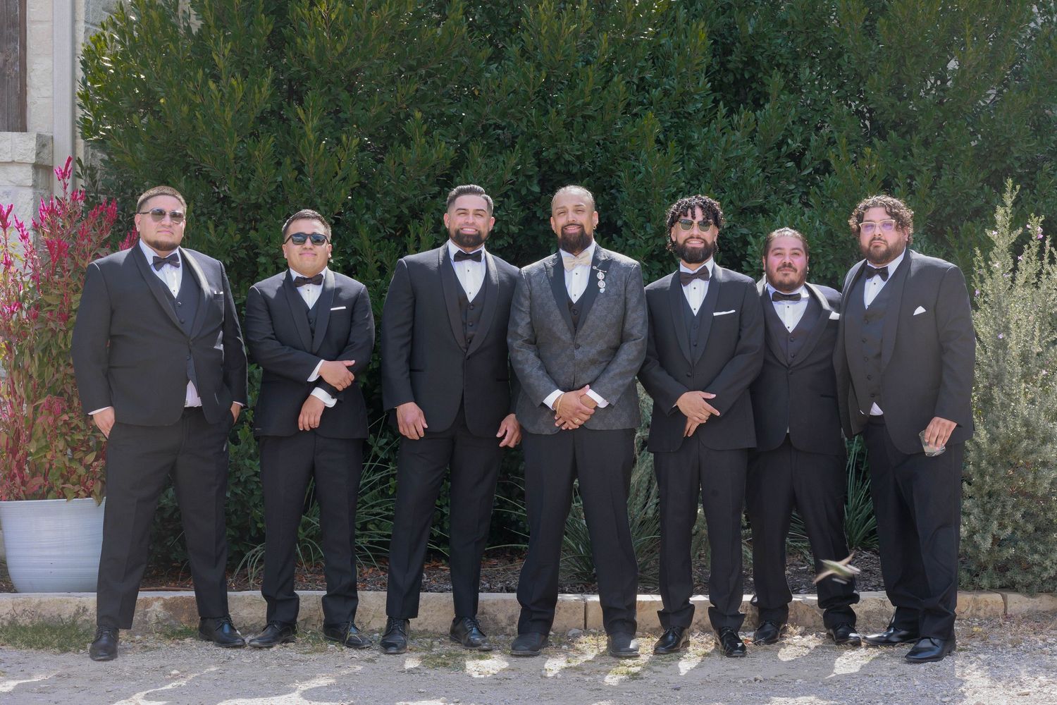 Groom and six groomsmen in charcoal suits posing outdoors before a wedding ceremony.
