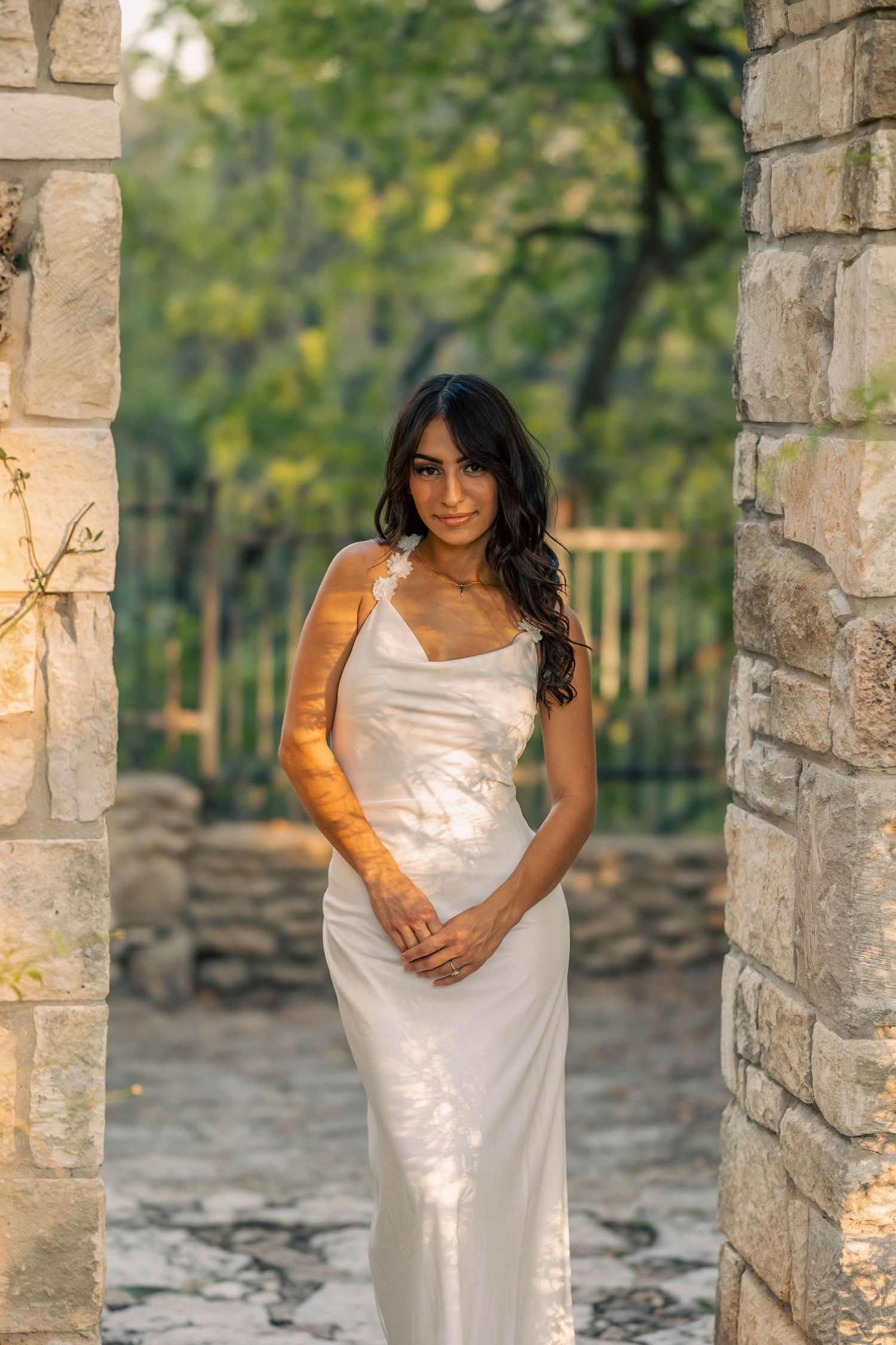 Bride in white slip dress standing between stone pillars in a garden at golden hour.