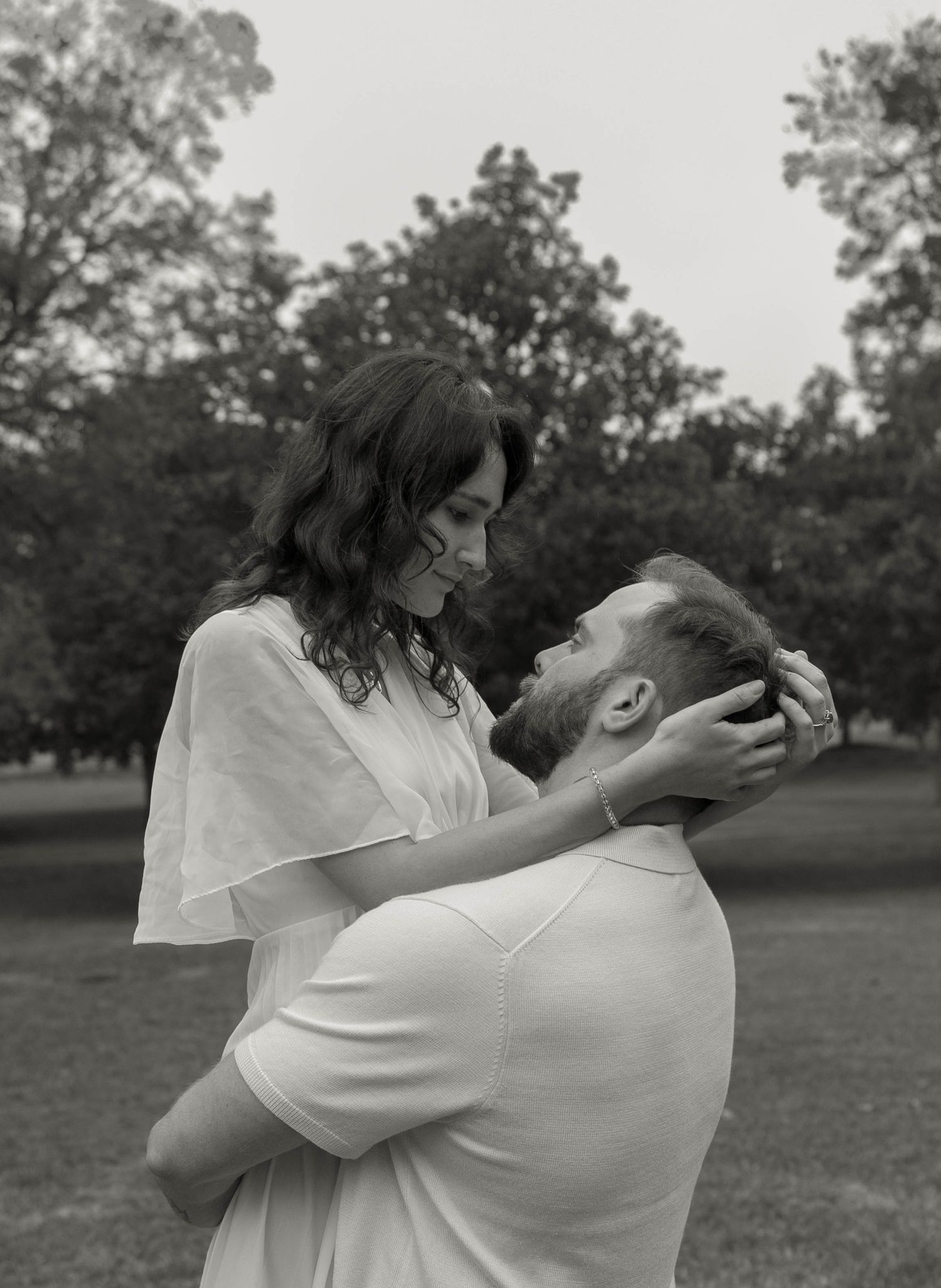 Black and white romantic photo of couple embracing in park, woman cradling mans face looking down at him.