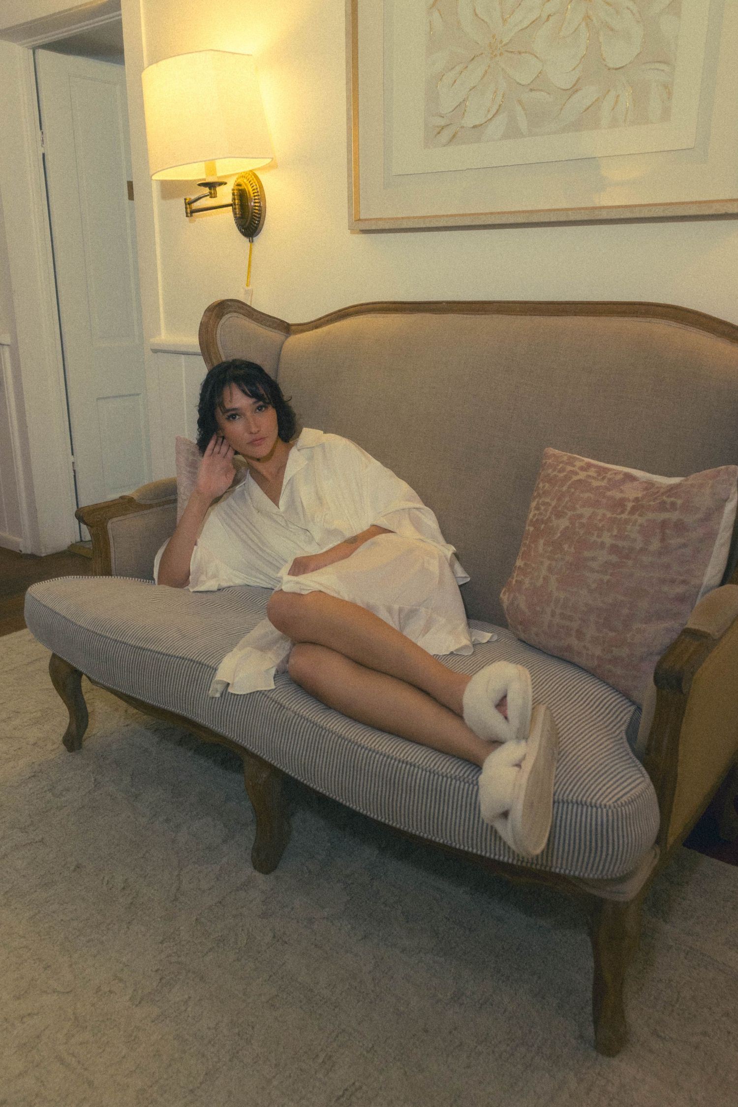 Bride relaxing on elegant sofa in white robe with fluffy slippers in warmly lit hotel room.