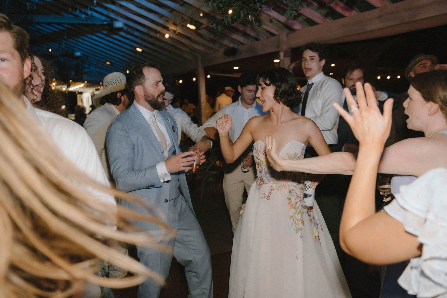 Bride and groom dance energetically with guests on crowded dance floor at lively indoor wedding reception.