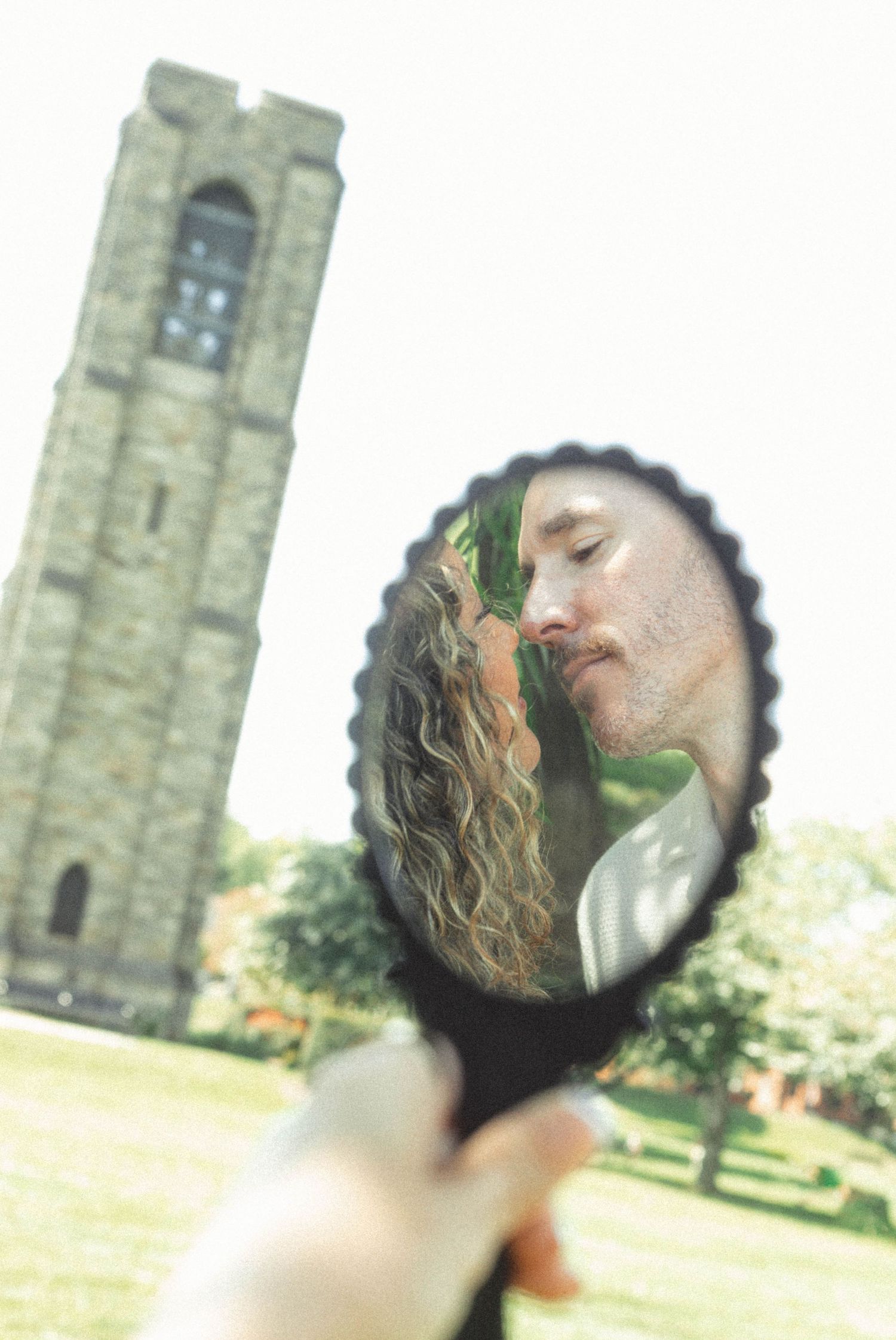A couple kissing reflected in a decorative hand mirror held up outdoors near a stone tower.