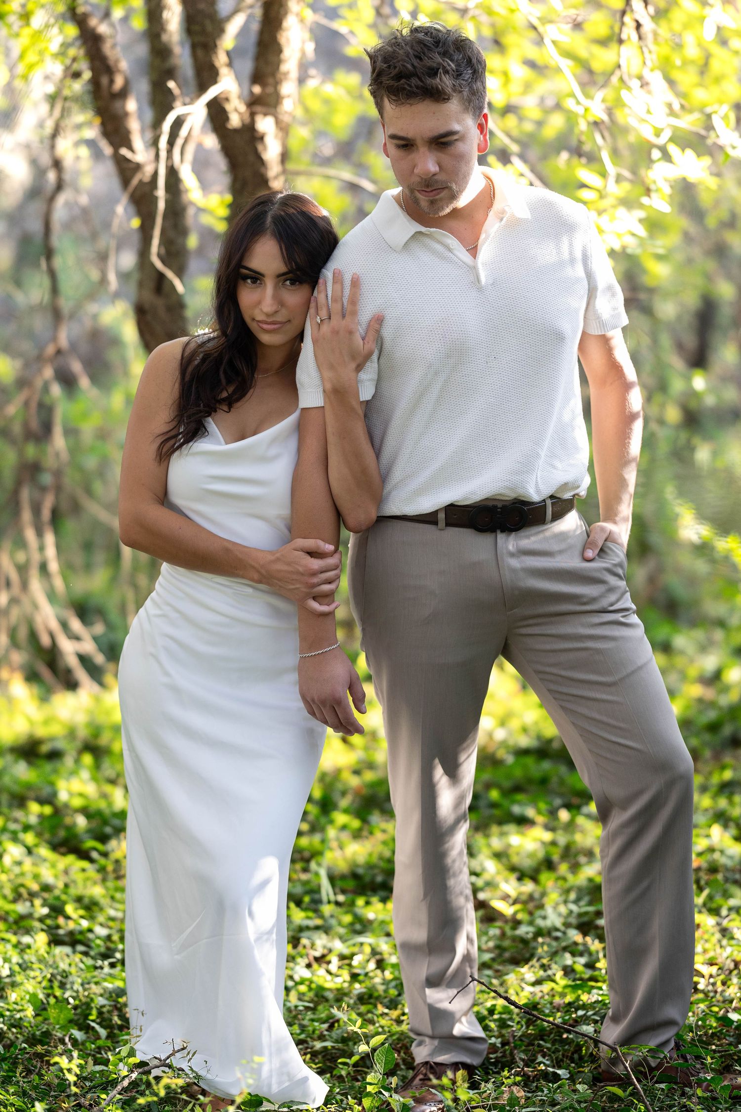 Elegant couple posing in sunlit forest, woman in white slip dress with man.
