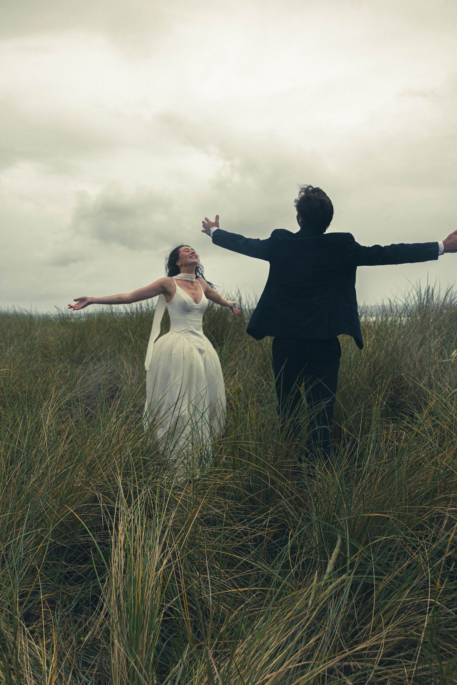 Silhouetted couple with outstretched arms stand together in tall grass field under moody cloudy sky.
