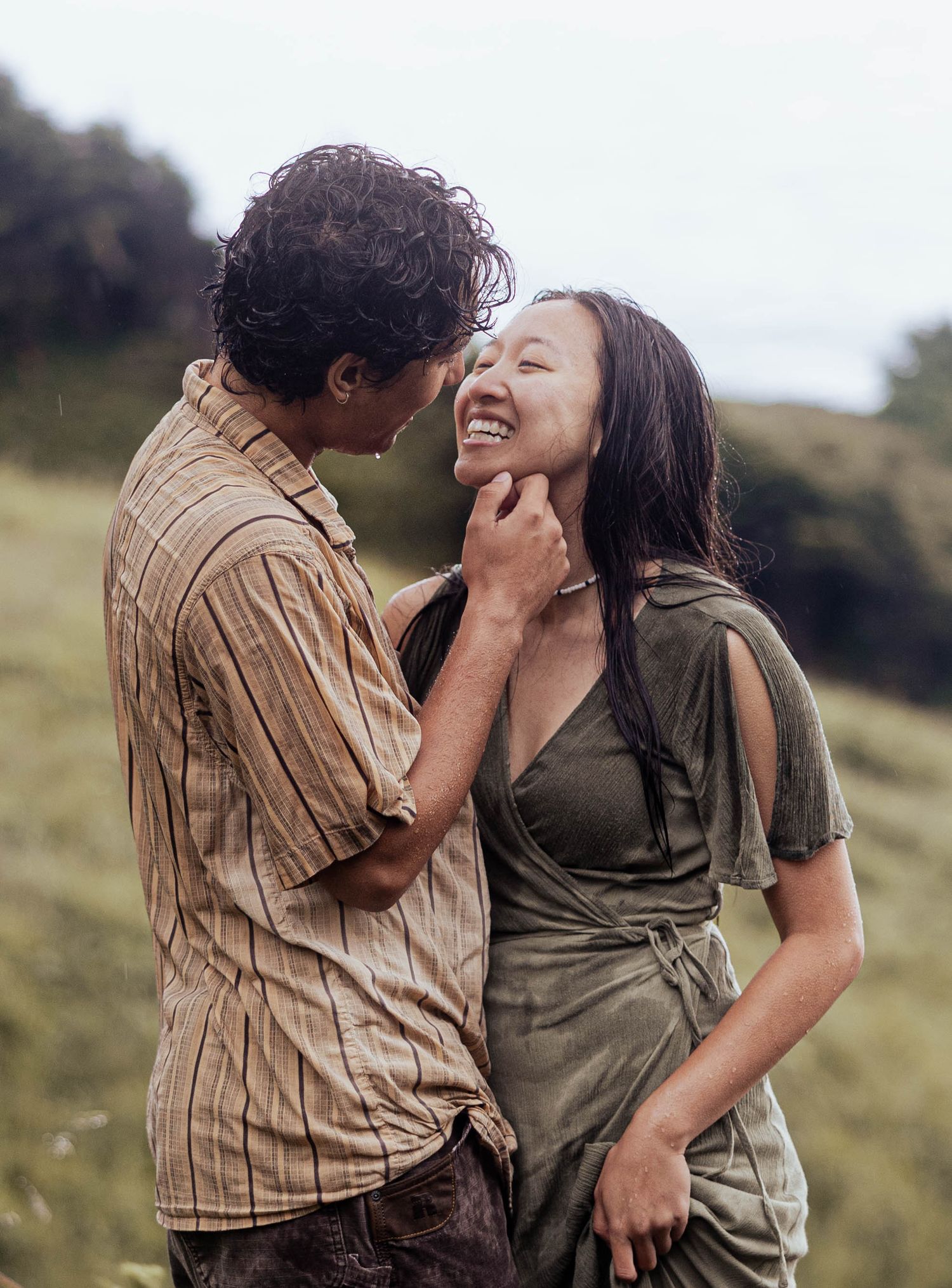 Happy Asian couple sharing joyful laugh in green meadow with ocean views in background.