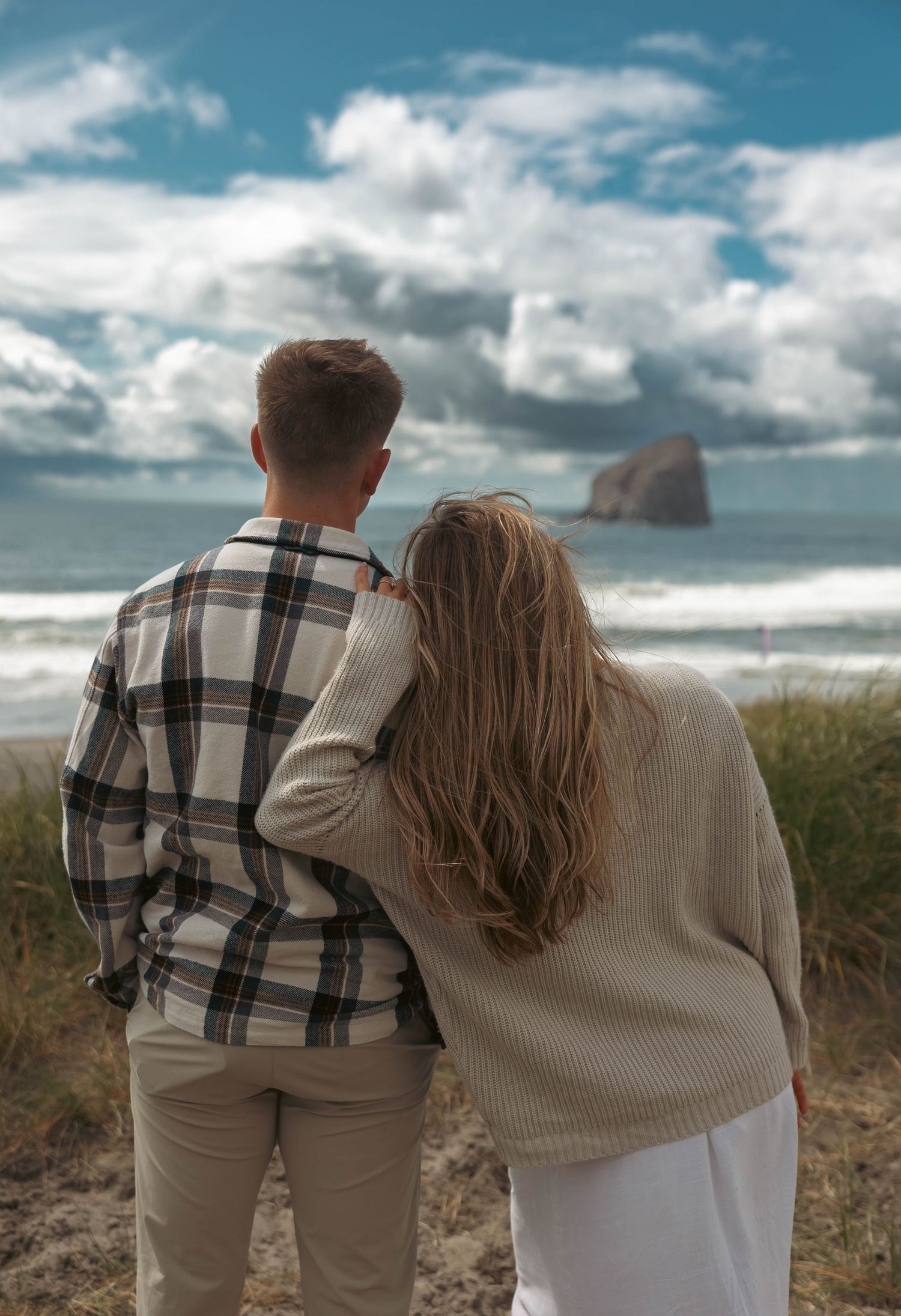 Couple standing together from behind, overlooking a scenic ocean view with dramatic clouds and a sea stack.
