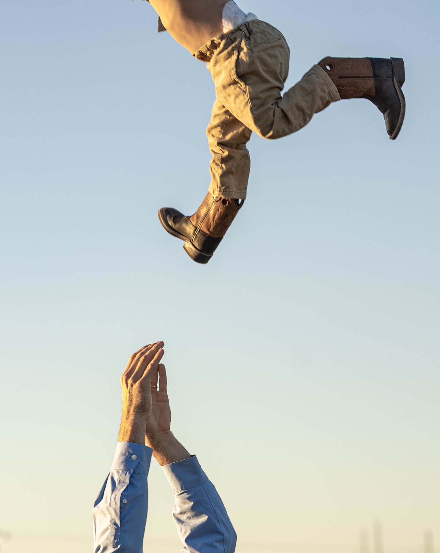 Child being tossed into the air by an adult with hands raised against a clear blue sky, wearing boots and khaki pants.