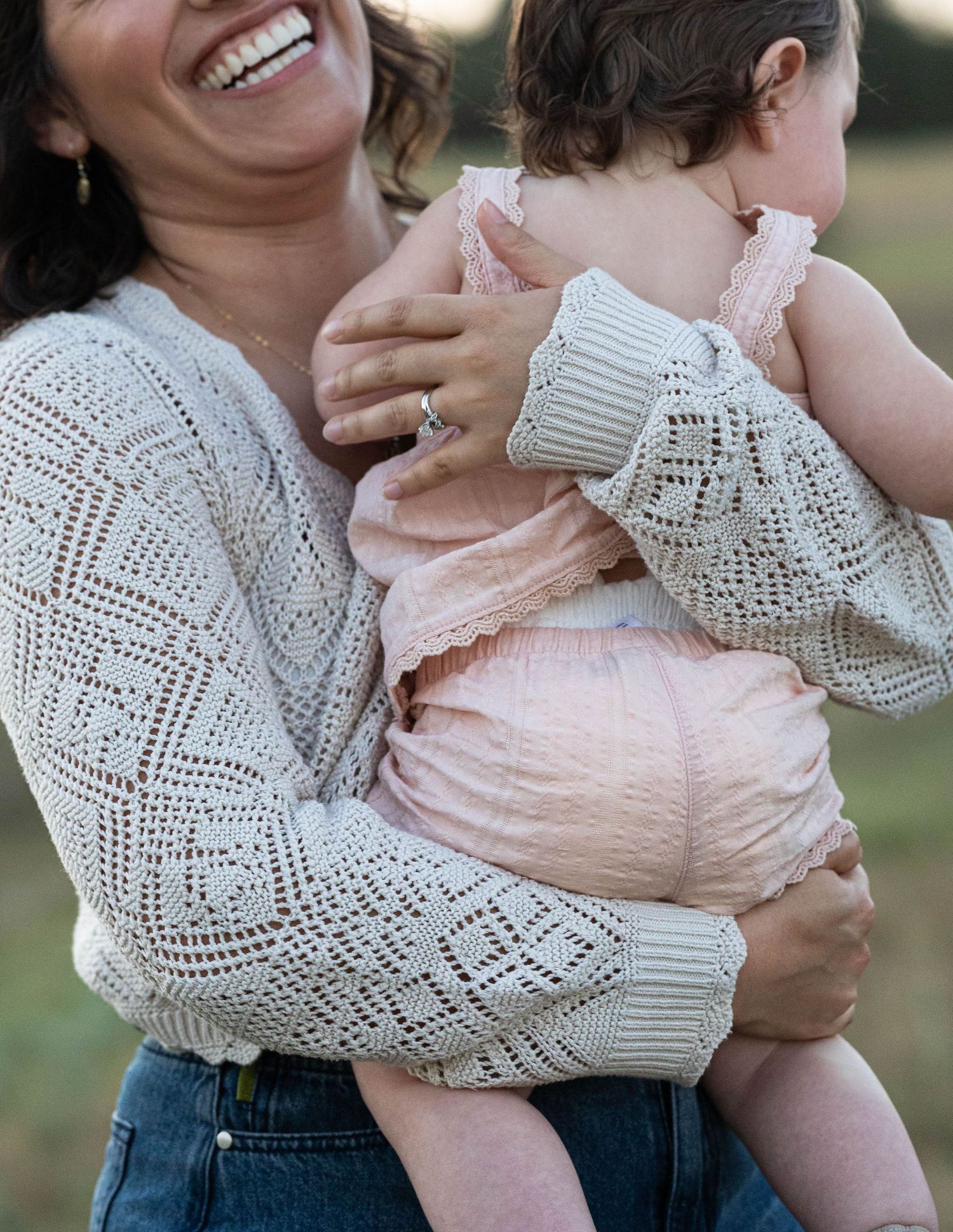 A laughing mother in a cream lace sweater lovingly holds her baby dressed in a pink outfit outdoors.