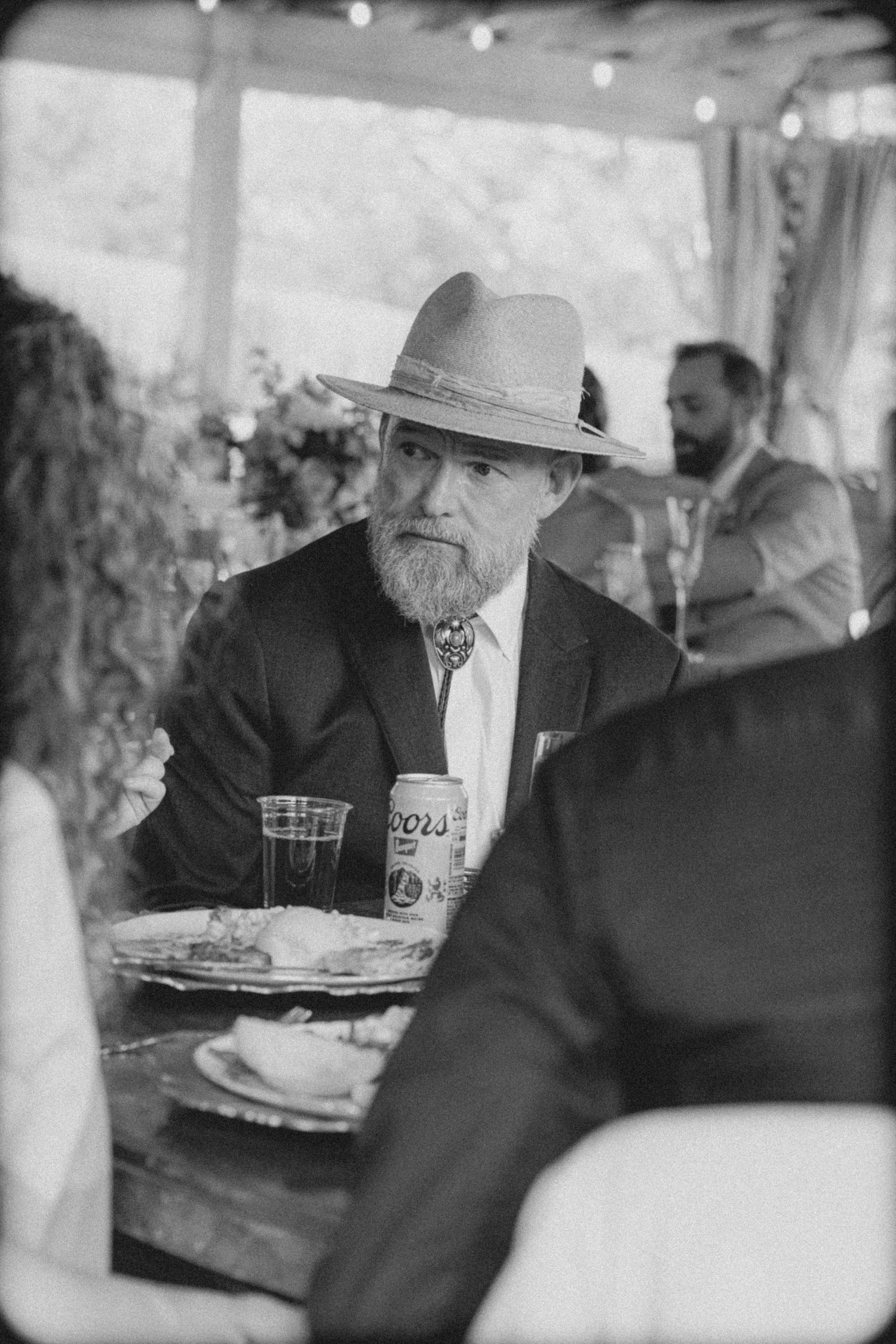 Older bearded man in a wide-brim hat and suit jacket sits at a wedding reception table with a Coors beer can.