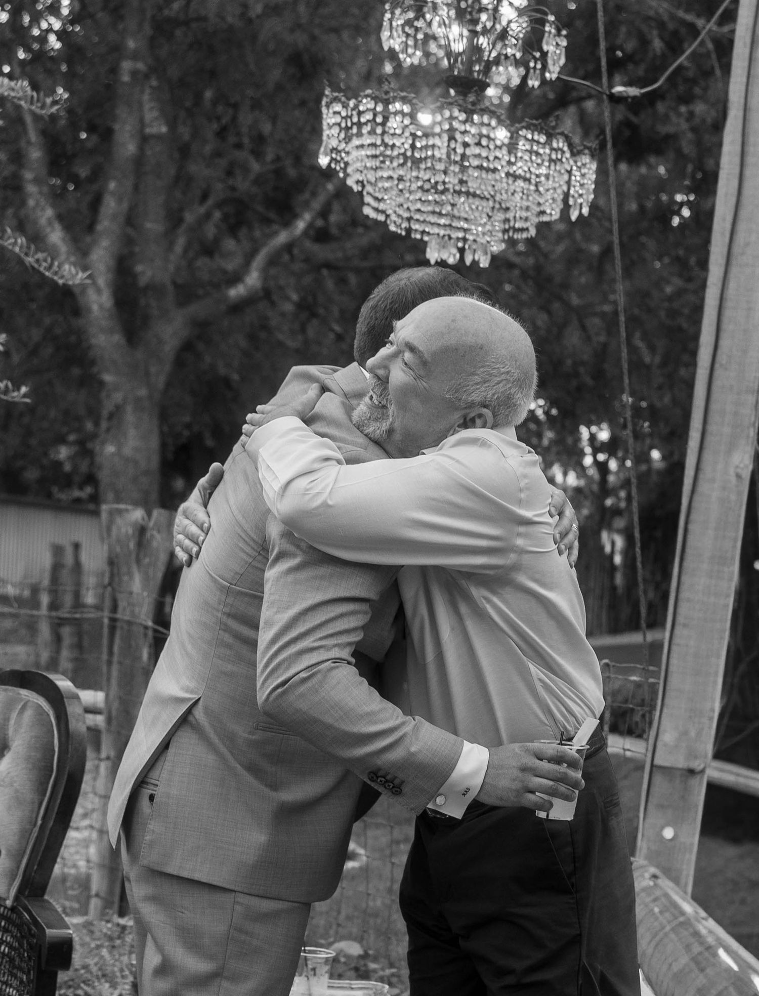 Two men sharing a warm embrace under chandelier at outdoor wedding in black and white photo.