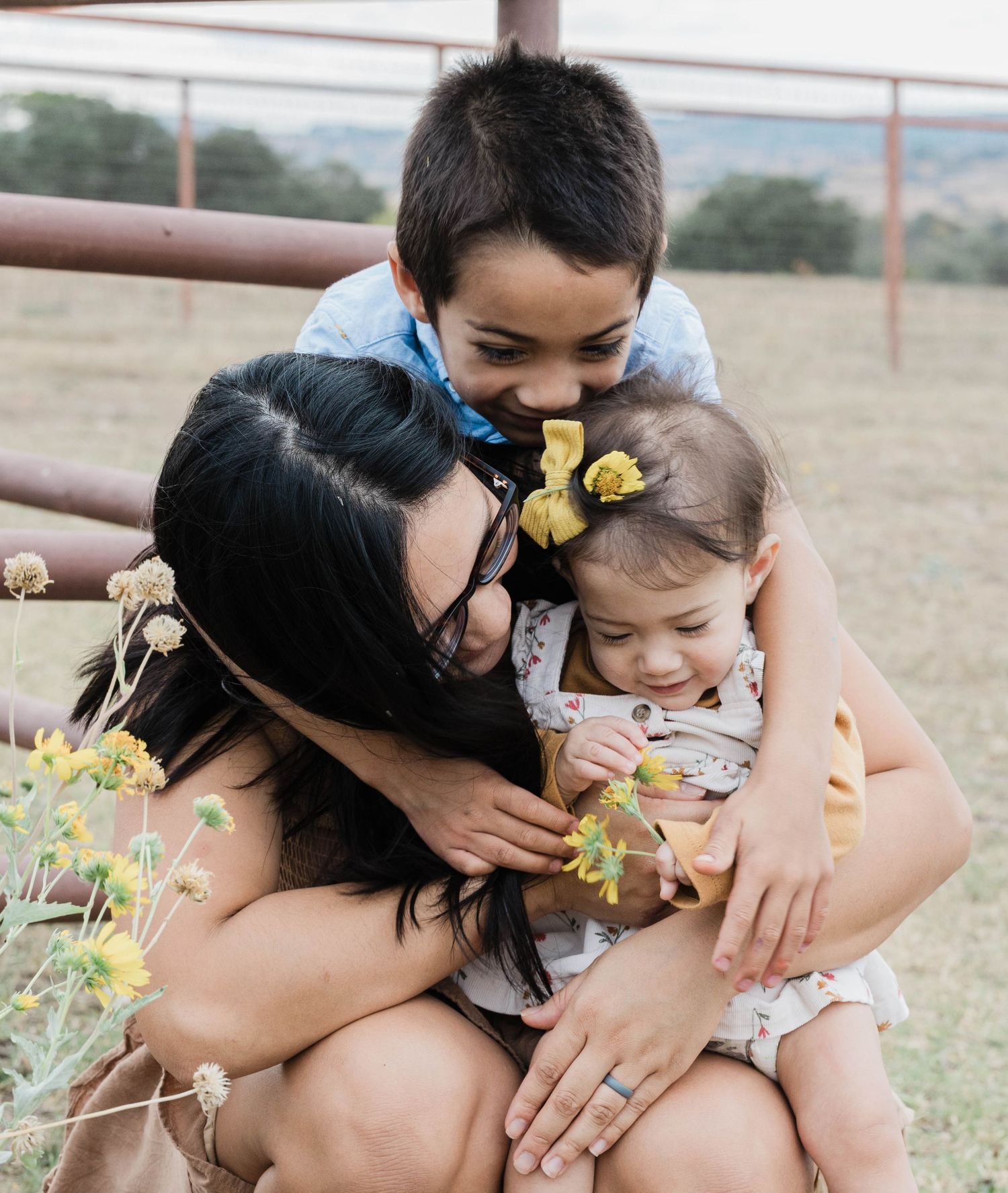 A loving mother crouches with her young son and baby daughter with yellow bow in a rustic outdoor setting.