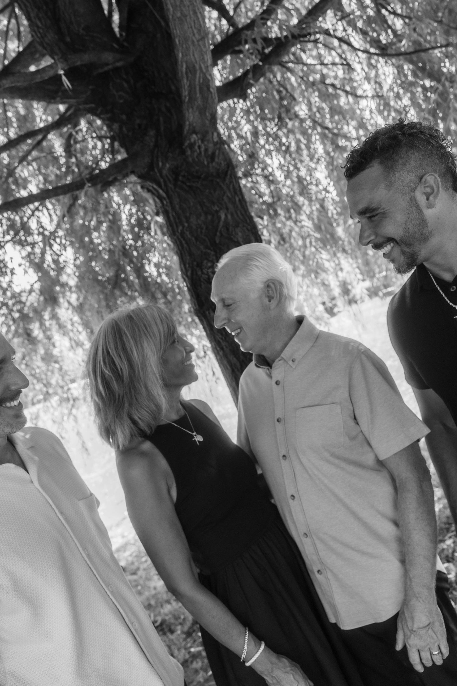 Black and white photo of an older couple smiling at each other under a tree, surrounded by family members.
