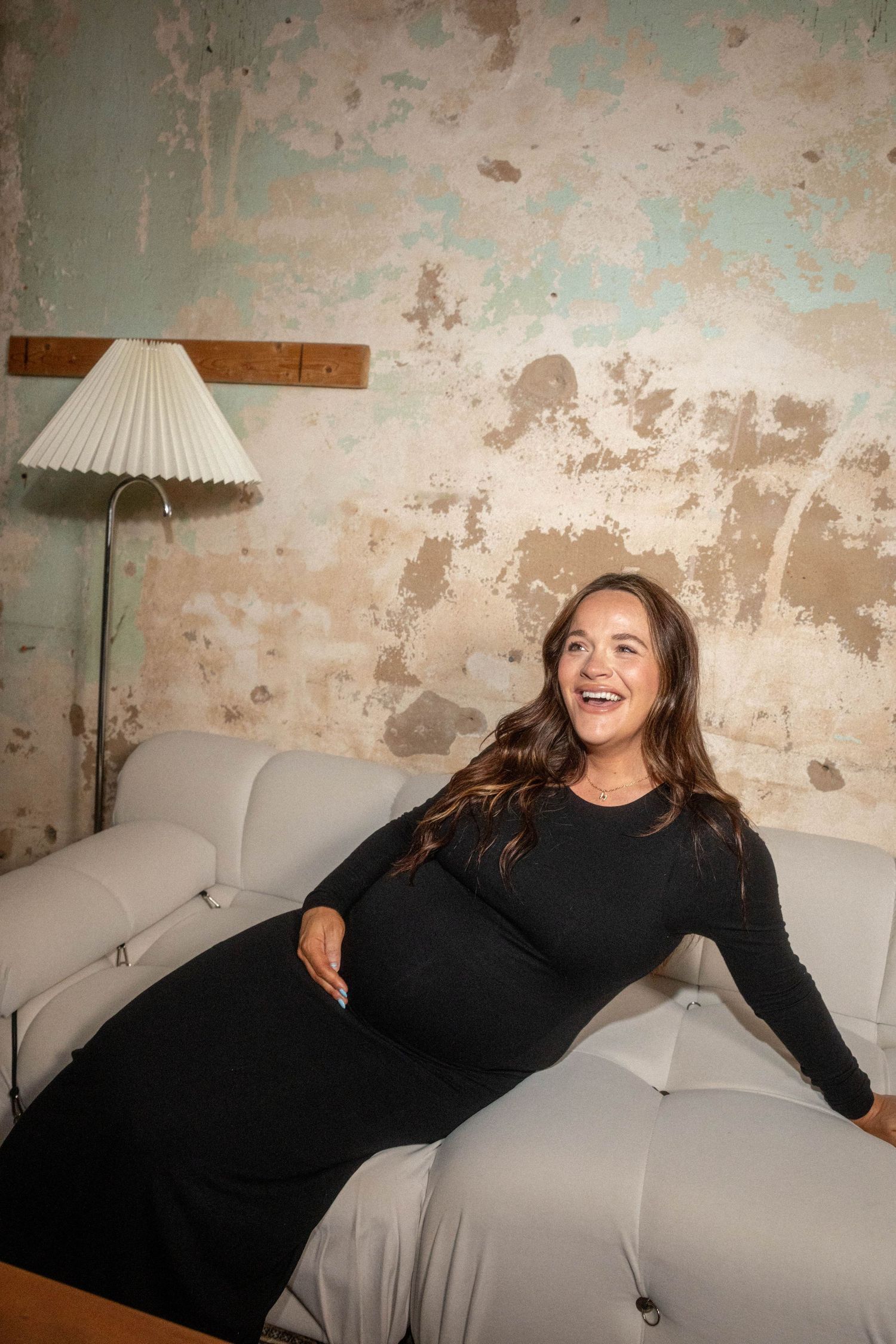 Laughing pregnant woman in black dress reclines on white sofa against rustic distressed wall with vintage lamp.