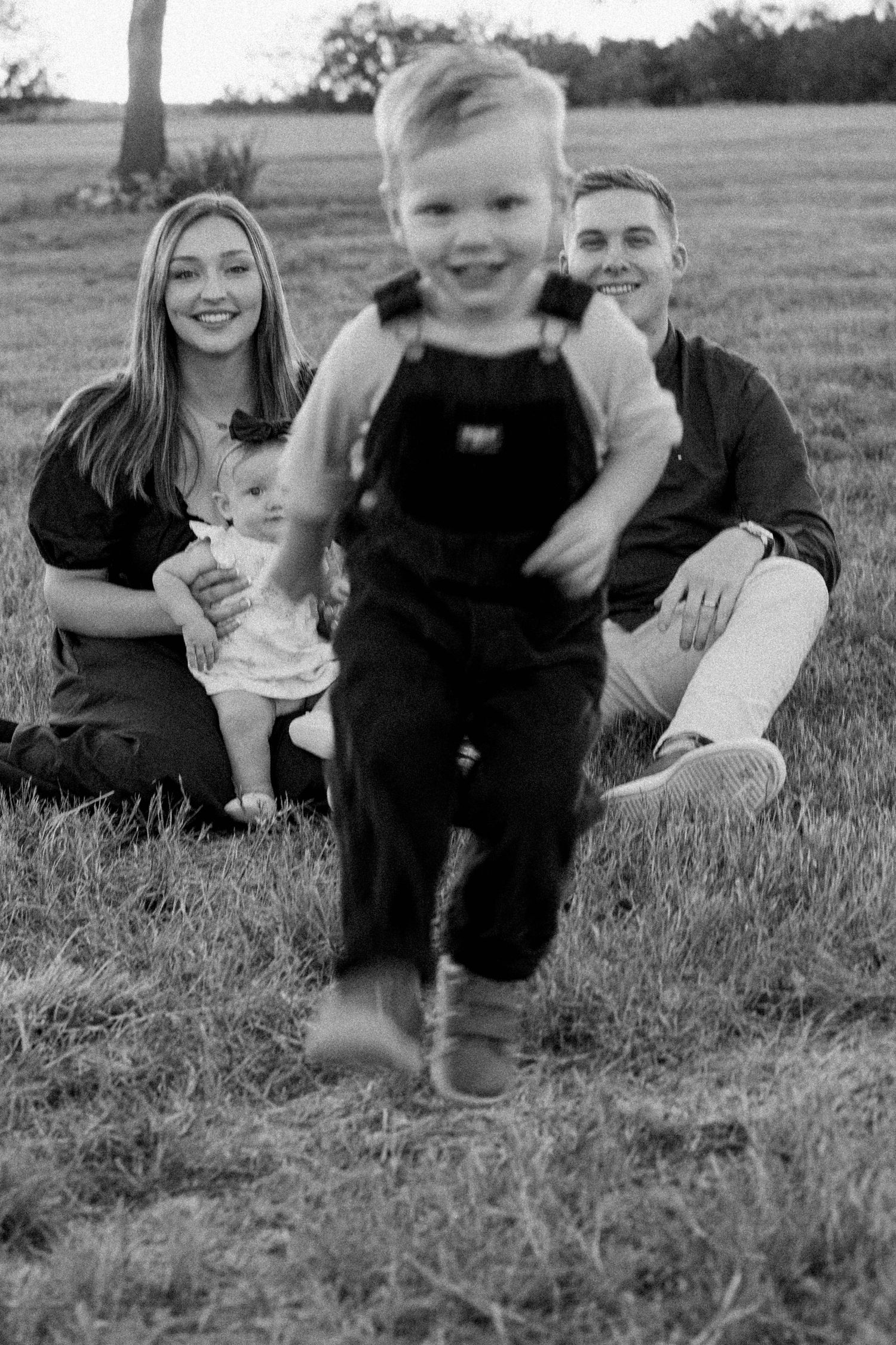 Black and white photo of happy toddler boy running in overalls toward camera with parents and baby sister in background.