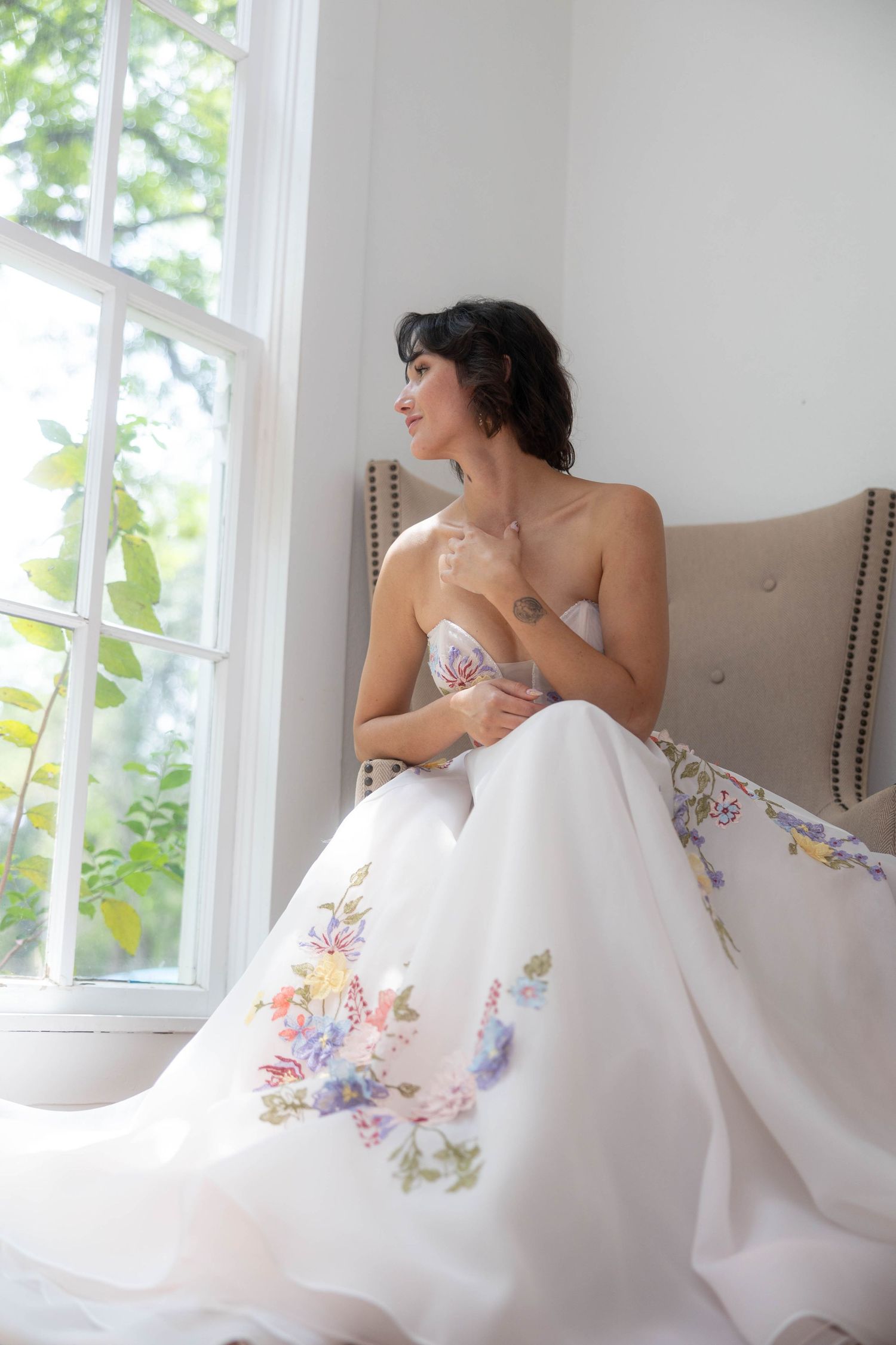 Bride in a strapless floral embroidered wedding gown seated by a sunlit window.