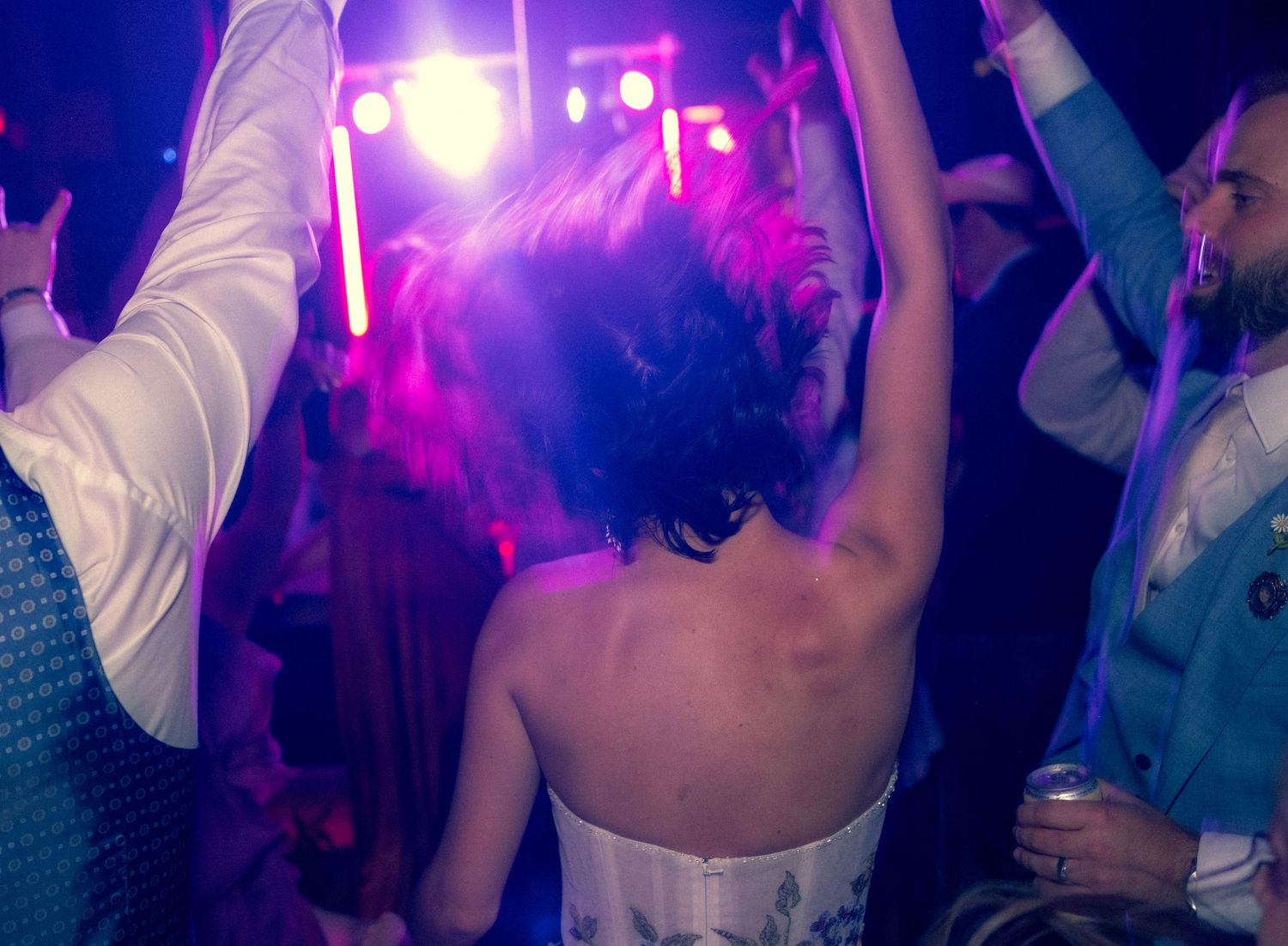 Bride with bare back dances energetically on colorfully lit wedding reception dance floor.