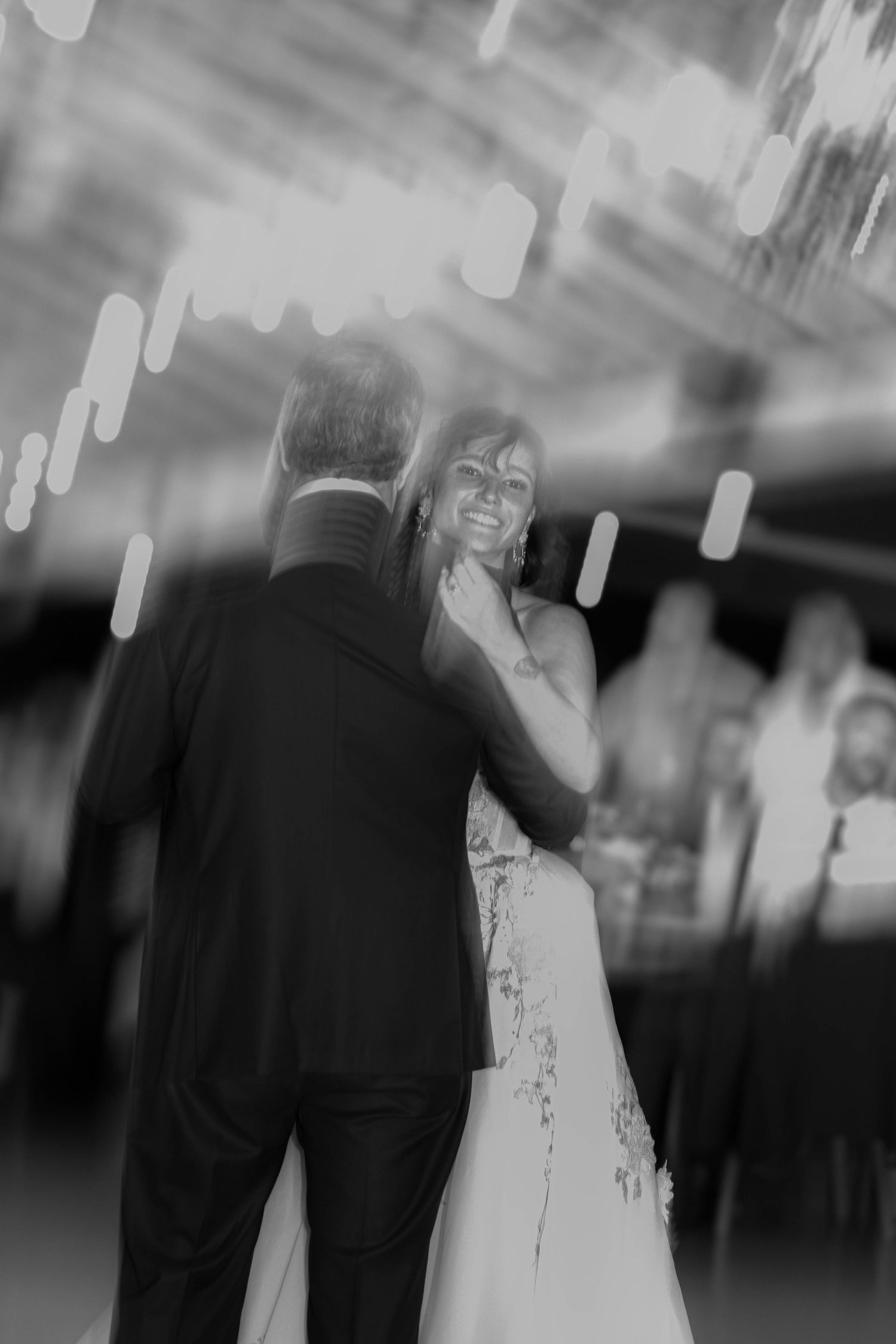 Bride smiles radiantly at groom during their first dance under glowing lights in a dreamy blurred black and white photo.