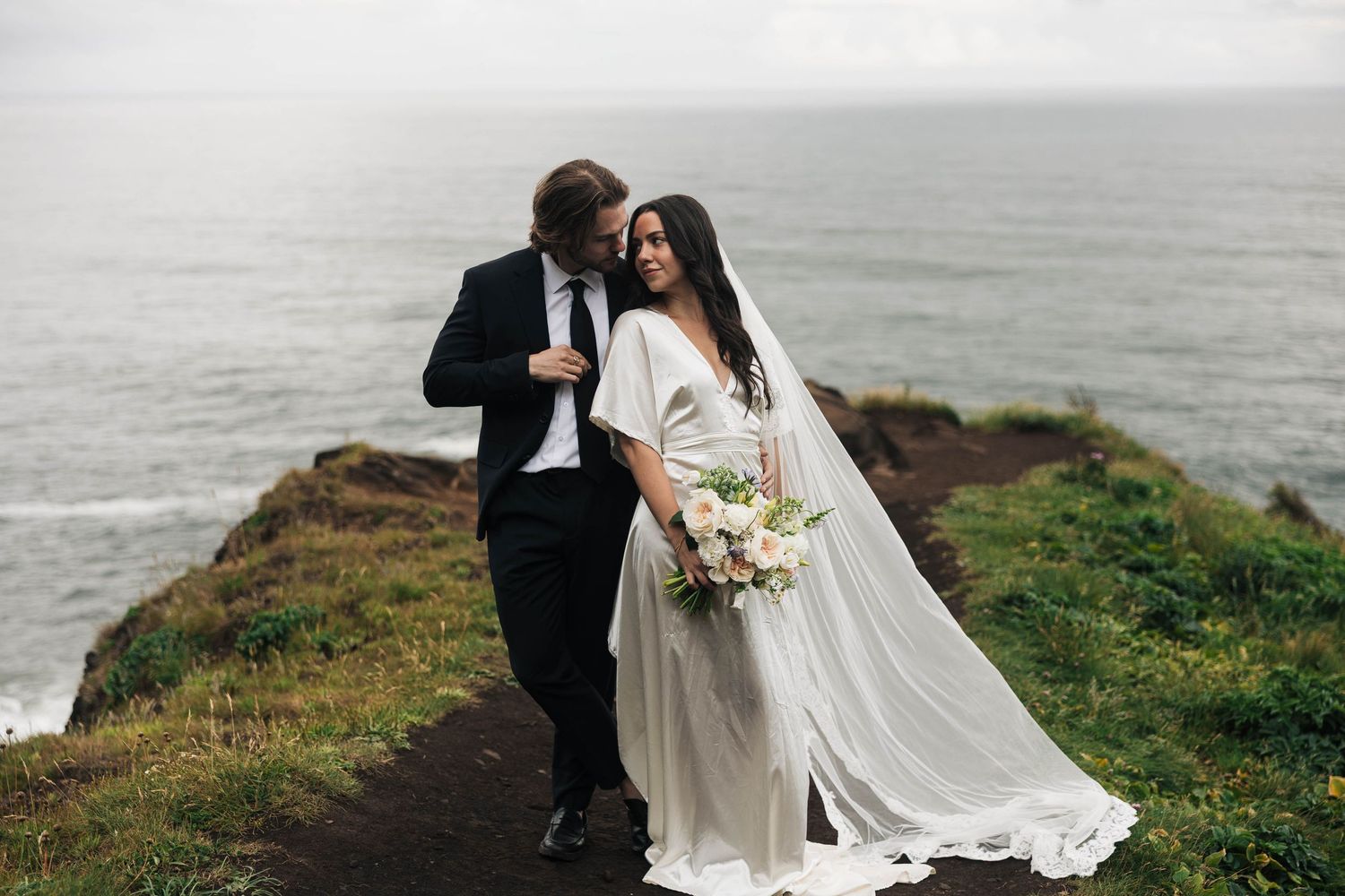Bride and groom share a romantic moment on a coastal cliff, with ocean waves in the background.