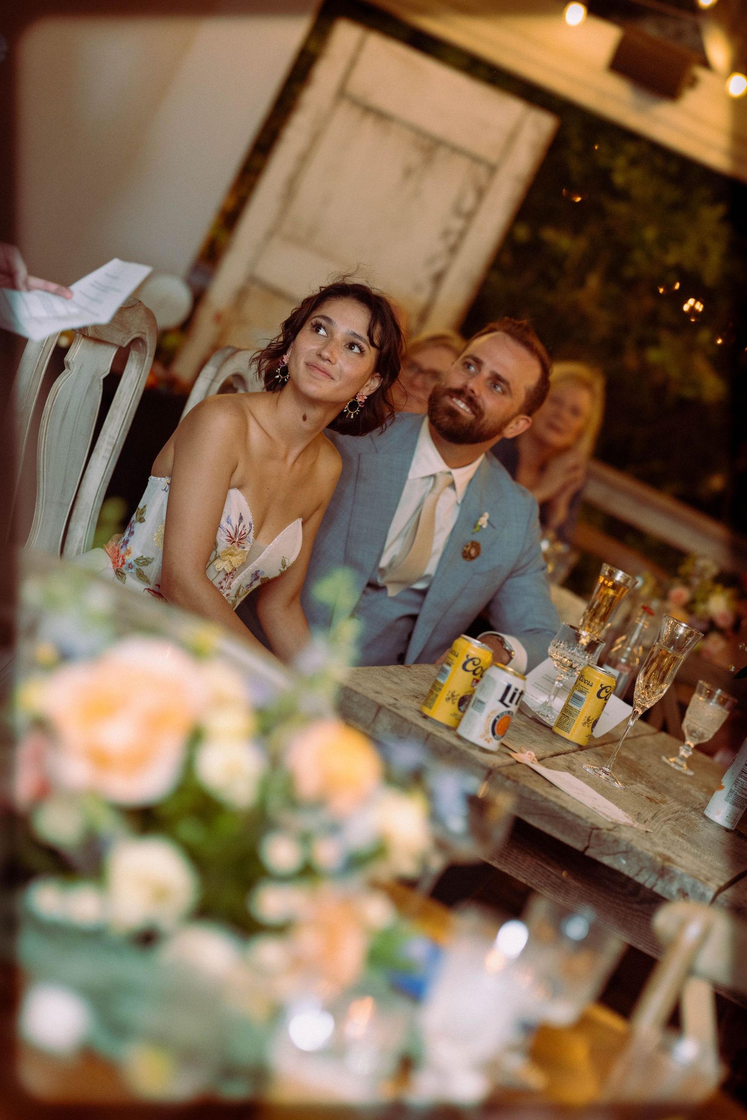 Bride in floral dress and bearded groom in gray suit listen attentively to speeches at their outdoor wedding reception.