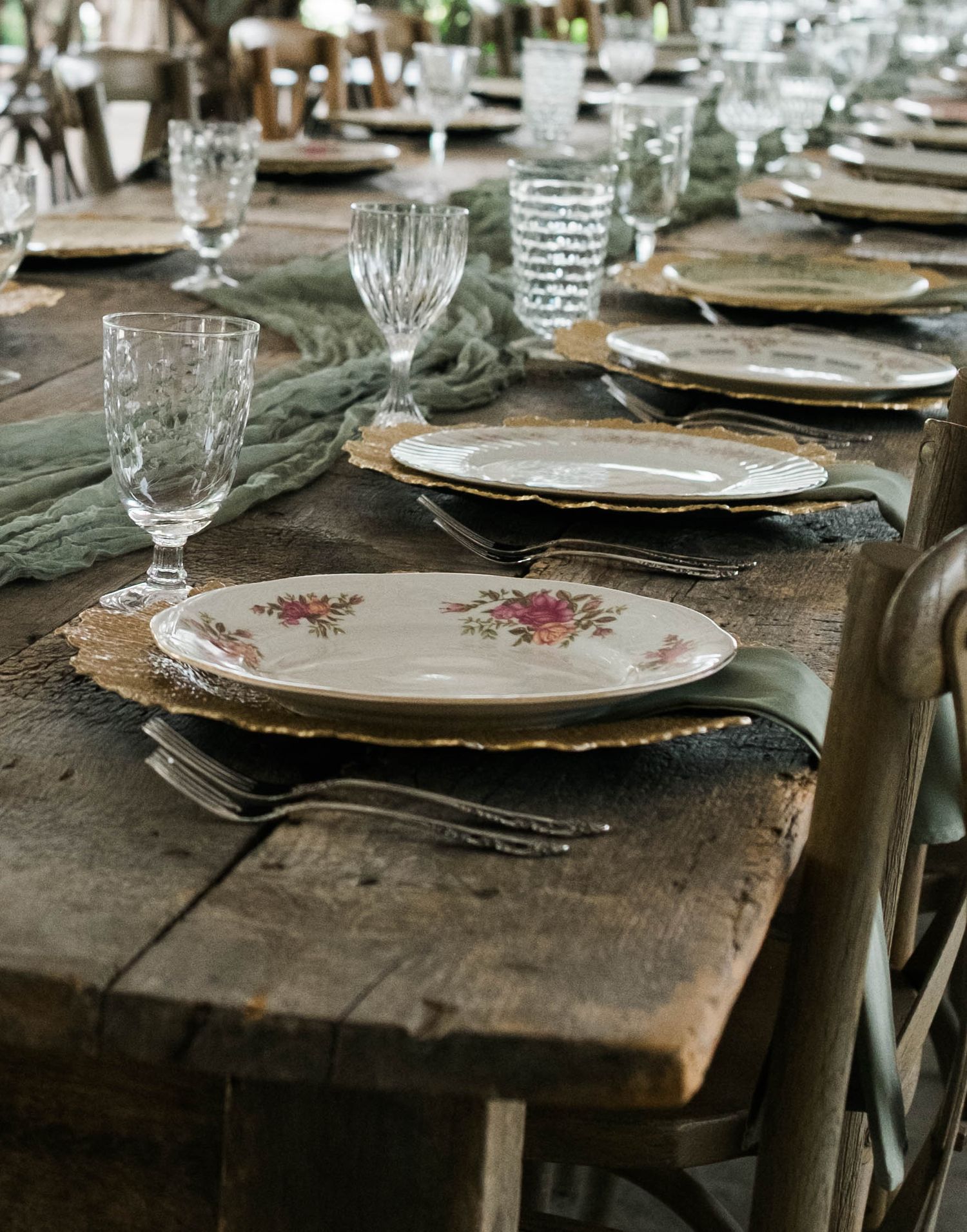 Rustic wooden wedding reception table set with vintage floral plates and crystal glassware.