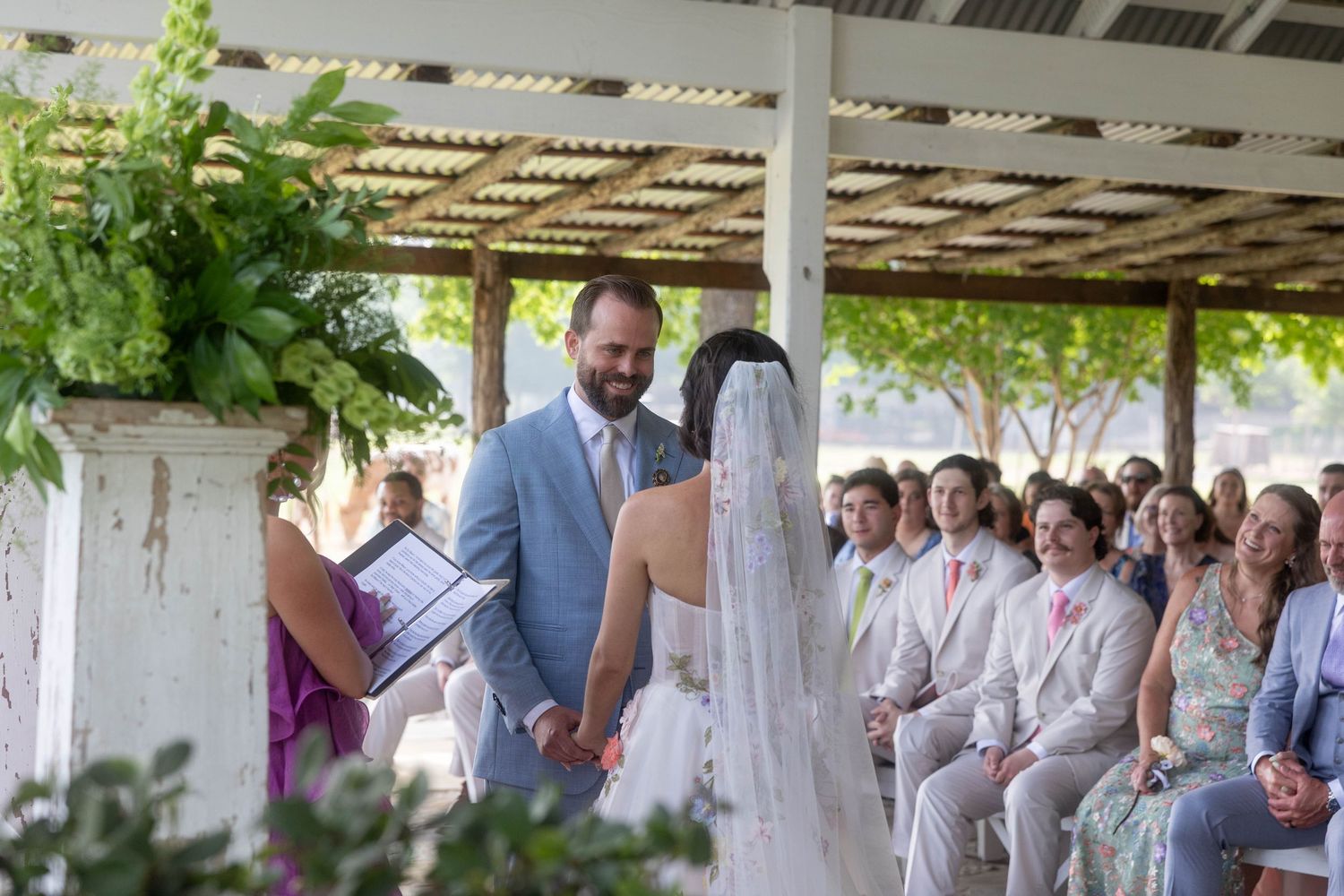 Groom holds brides hands during outdoor wedding ceremony under a rustic pavilion.