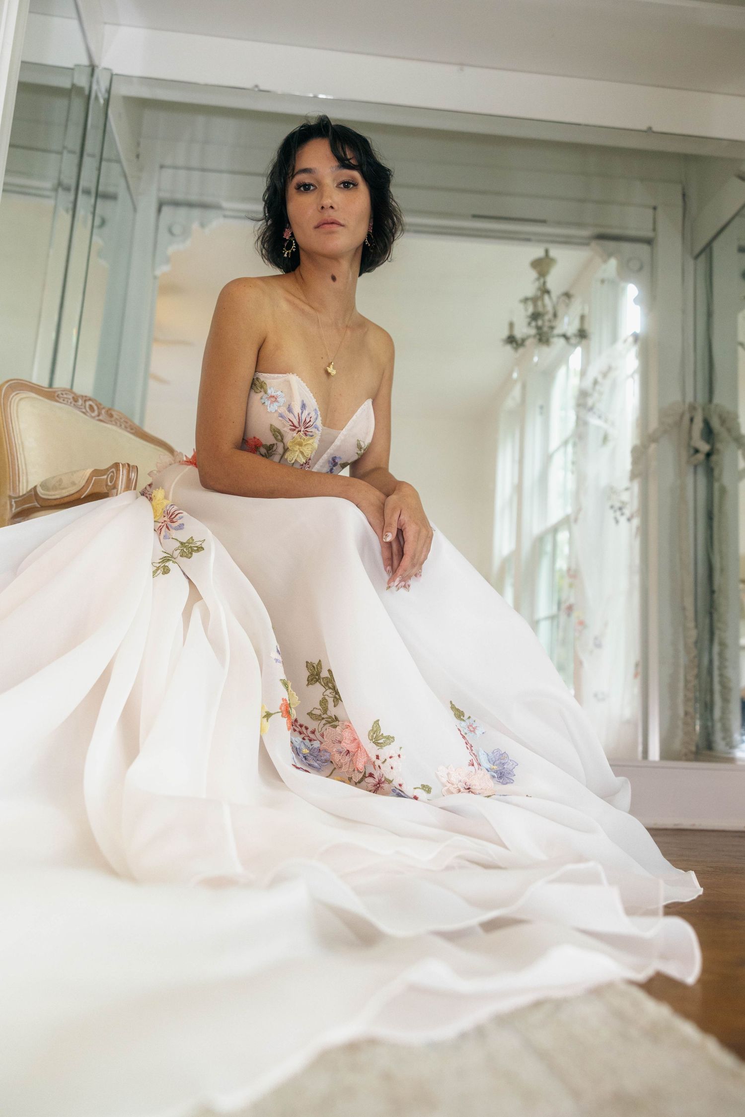 A bride in a strapless floral ball gown with a long flowing train poses seated in a bridal boutique.