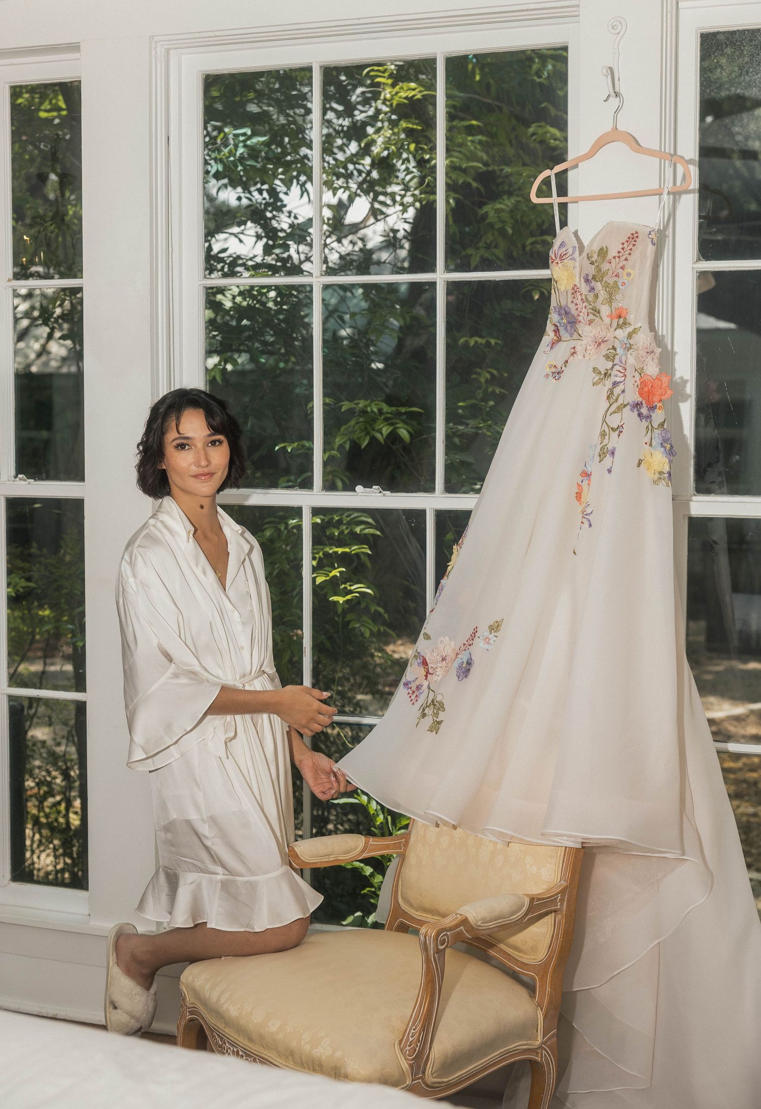 Bride in silk robe admires her floral embroidered wedding gown hanging by a sunlit window.