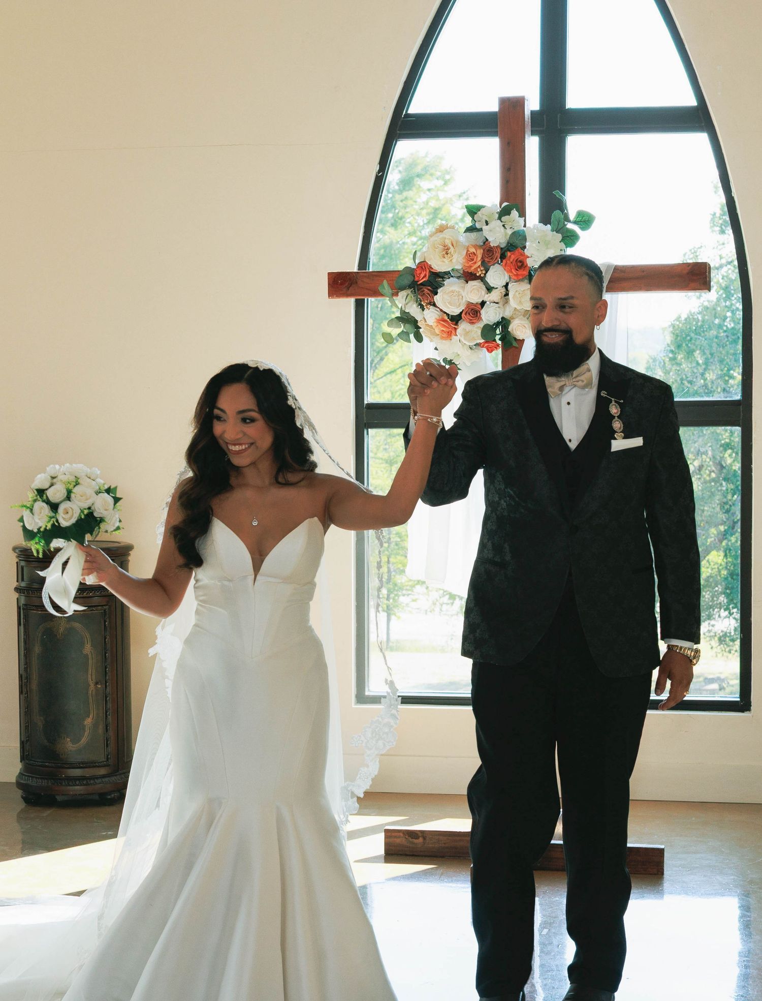 Bride and groom celebrate at church altar with floral cross backdrop, bride holding white bouquet.