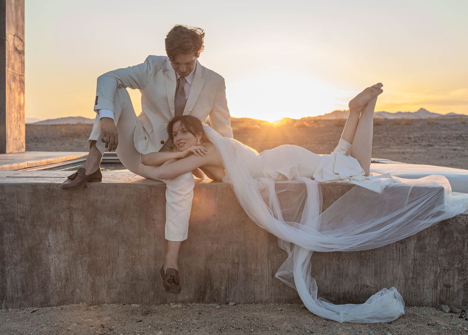 Bride and groom posing dramatically at sunset ruins, bride in flowing veil laying across concrete wall.