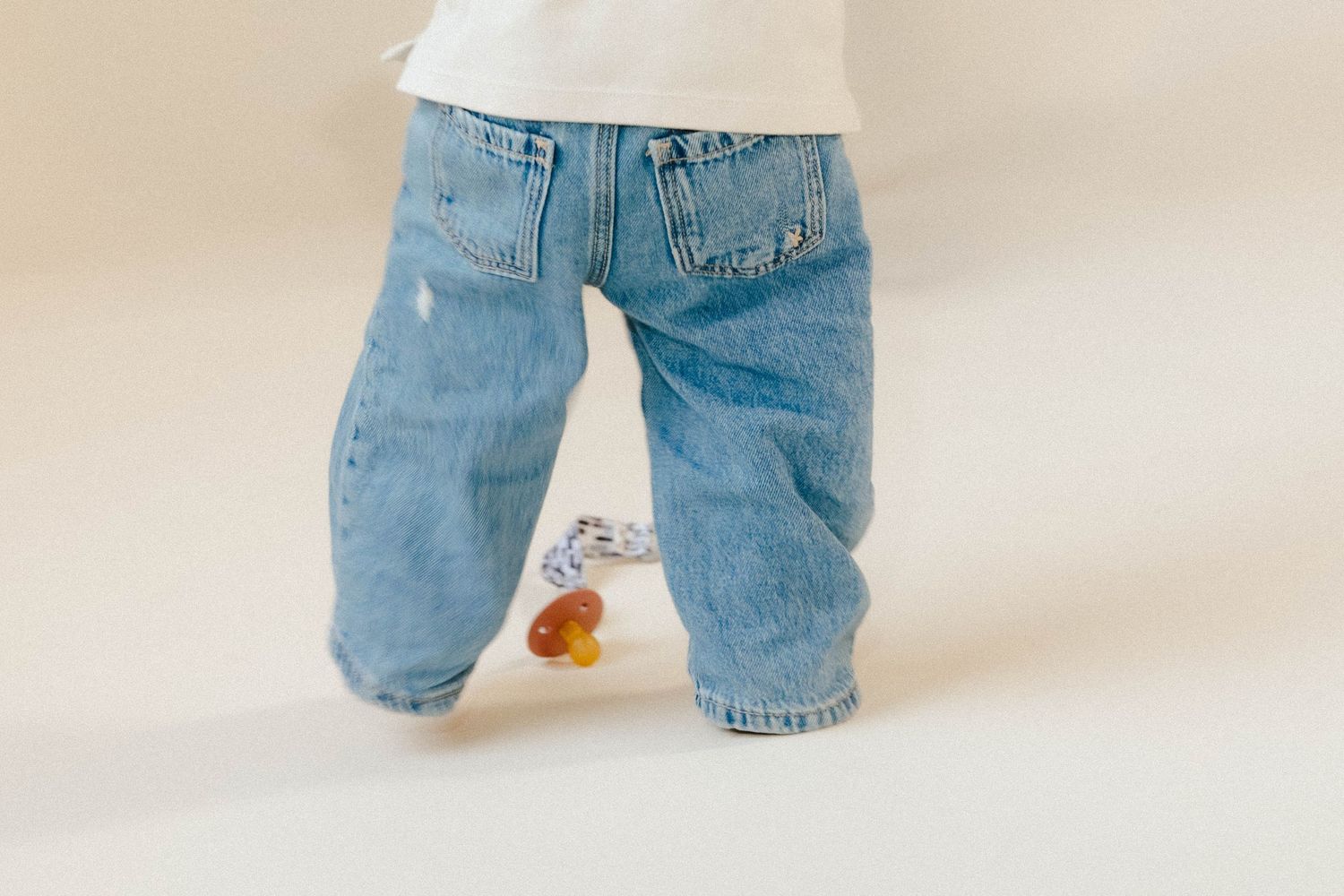 Close-up of a toddler in light blue jeans with a pacifier on the floor beside their feet.