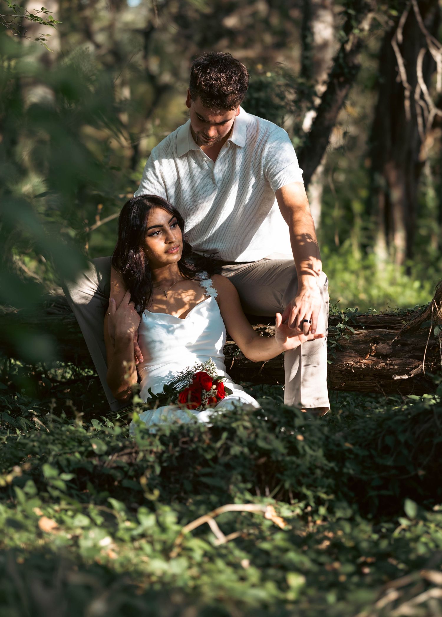 Couple sitting together in a sunlit forest with red roses nearby.