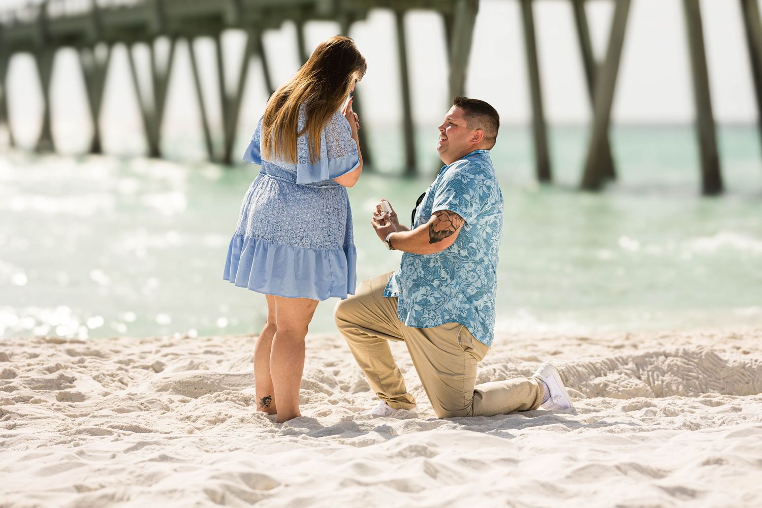 Man proposing on one knee to woman on sandy beach near pier with turquoise ocean.