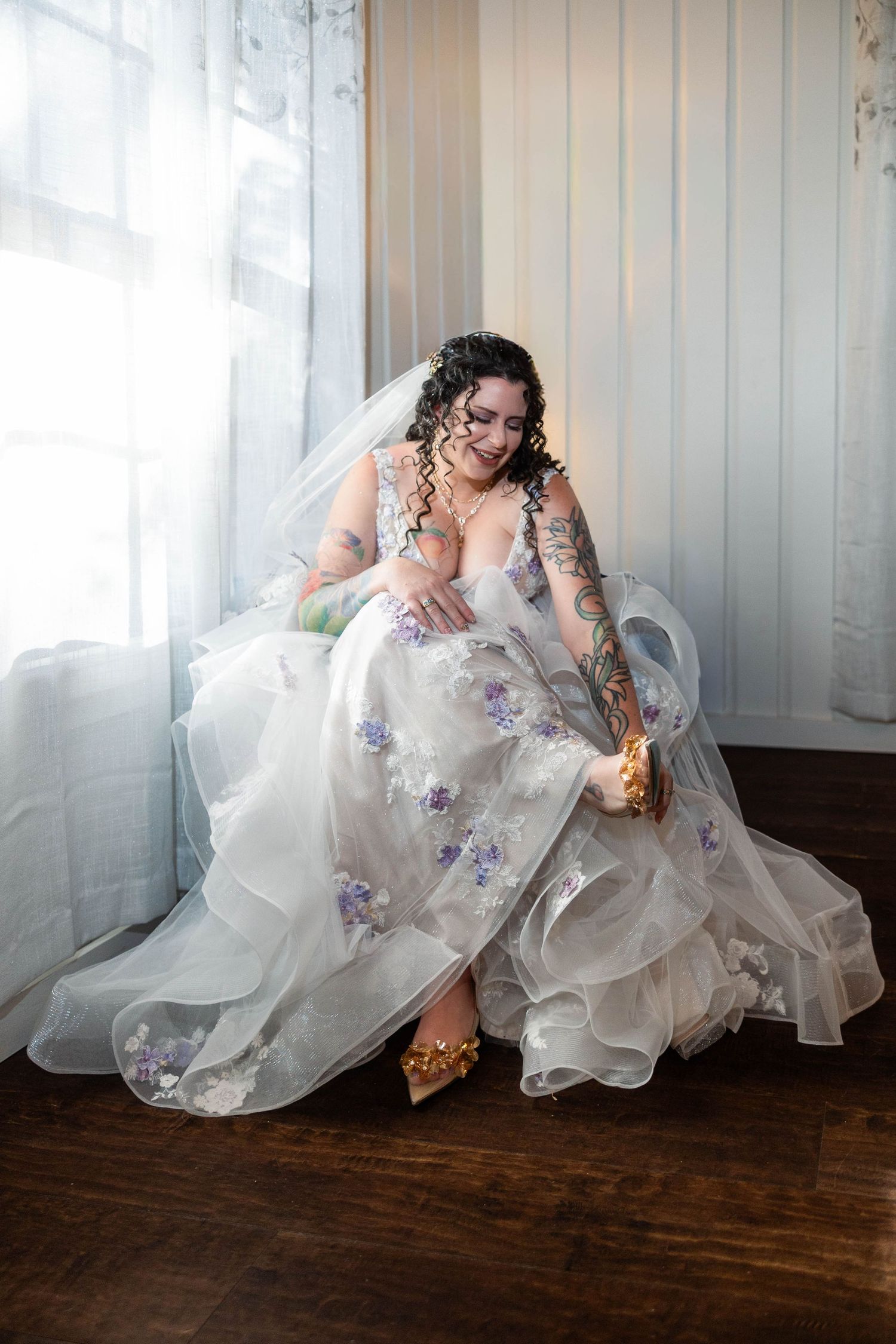 Tattooed bride in floral wedding gown with veil sitting by window putting on gold heels.