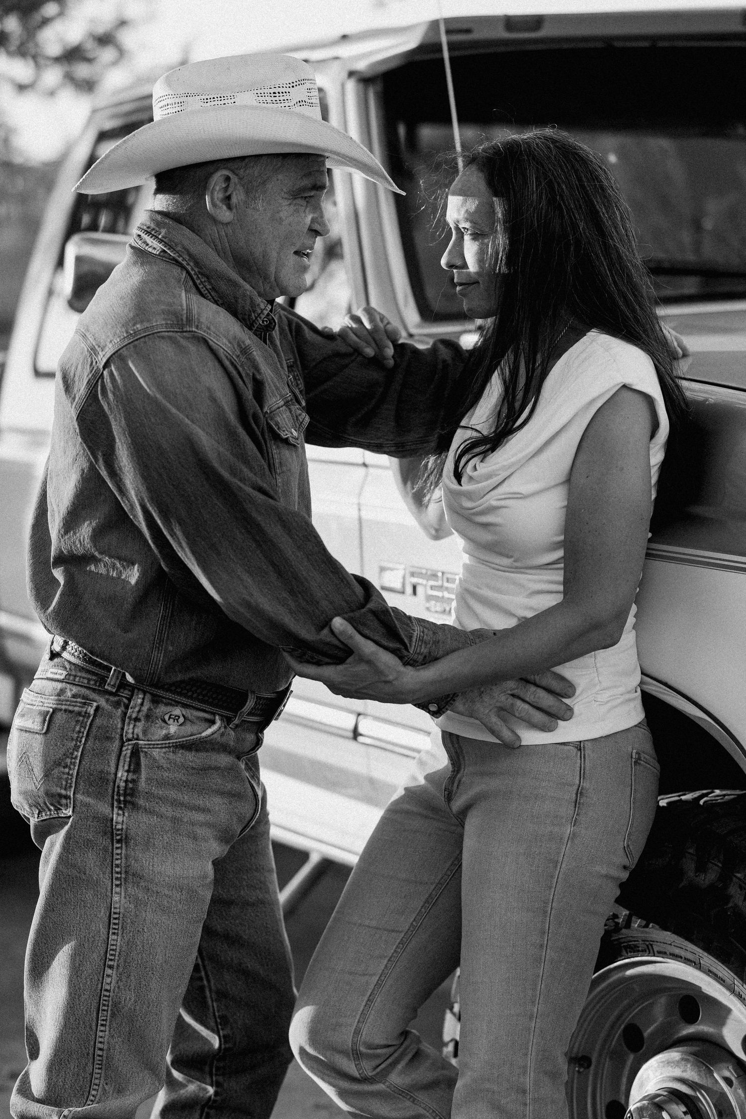 A cowboy in a white hat and denim shirt holds a dark-haired woman close near a truck in a black and white photo.