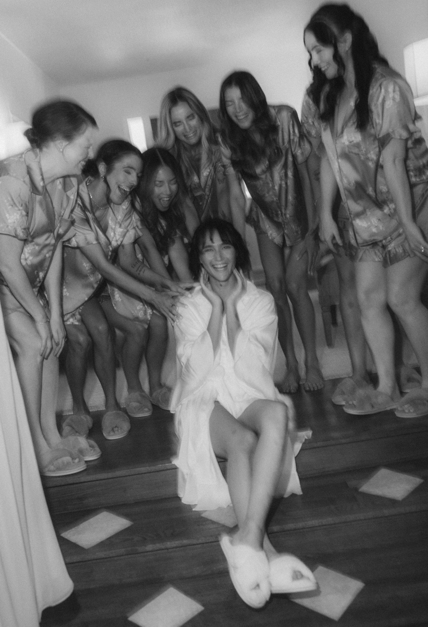 Black and white photo of a bride in a white robe surrounded by laughing bridesmaids in casual attire.